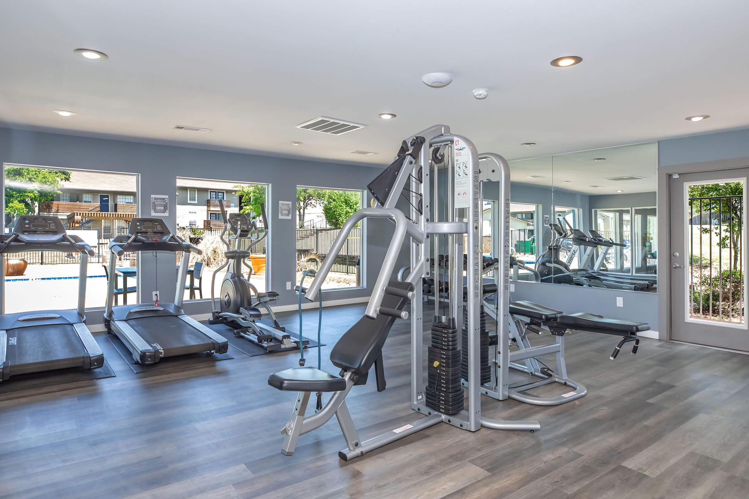 A modern gym interior featuring workout equipment, including a multi-station strength machine and treadmills. Large windows allow natural light to fill the space, and there are views of an outdoor area. The floor has a sleek wooden design, creating an inviting atmosphere for exercise.