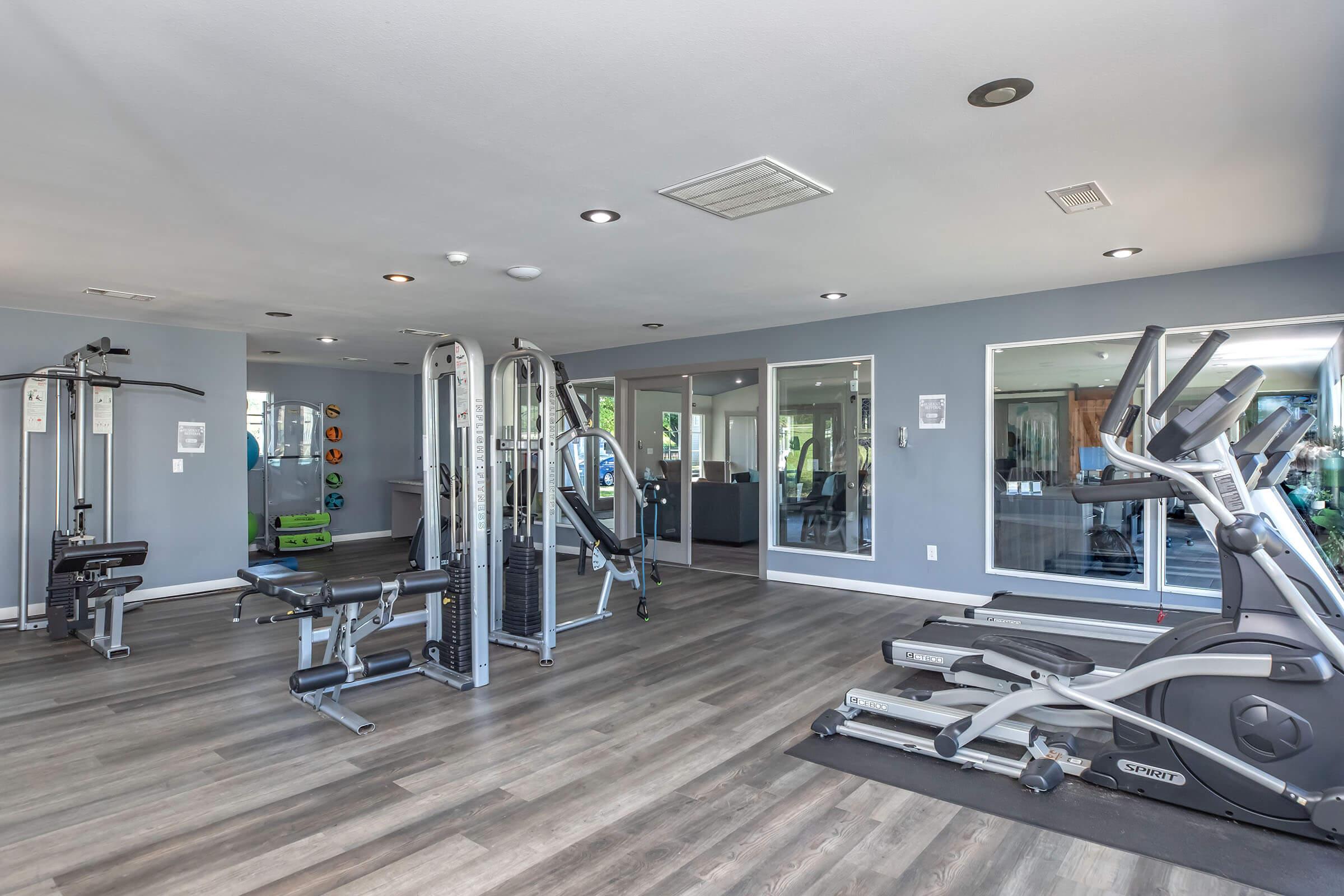 A modern gym featuring various fitness equipment, including weight machines, cardio machines like treadmills and ellipticals, and free weights. The well-lit space has a neutral color scheme with large windows allowing natural light. Mirrors on the walls enhance the open and inviting atmosphere.