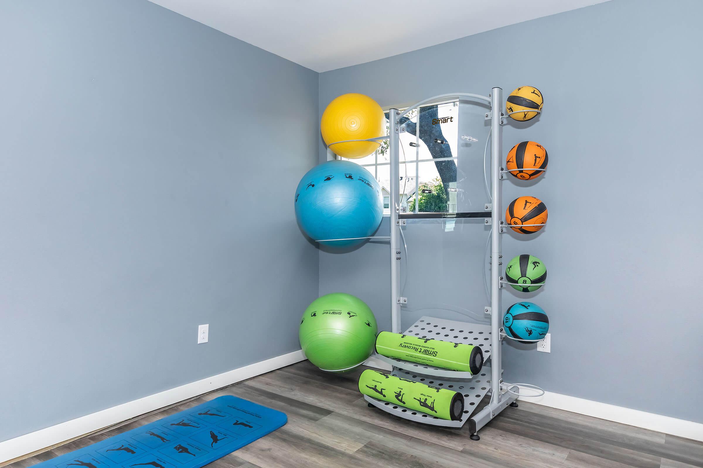 A fitness corner featuring exercise balls in various colors mounted on a wall, a mirrored equipment stand, and a green exercise mat on the floor. The walls are painted light blue and the flooring is wooden.