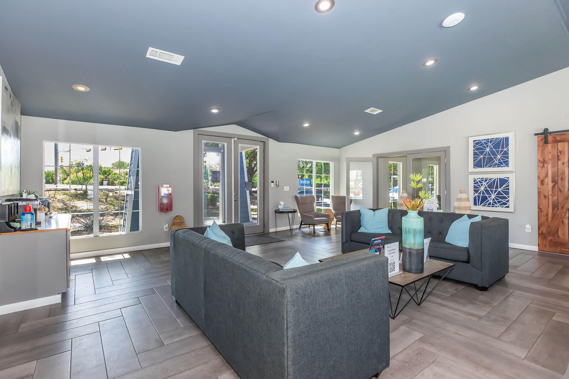 A modern lounge area featuring two grey sofas and a coffee table, with light blue decorative pillows. There are large windows letting in natural light, a sliding barn door, and decorative wall art. A small kitchenette with a water cooler and plants adds to the inviting atmosphere.