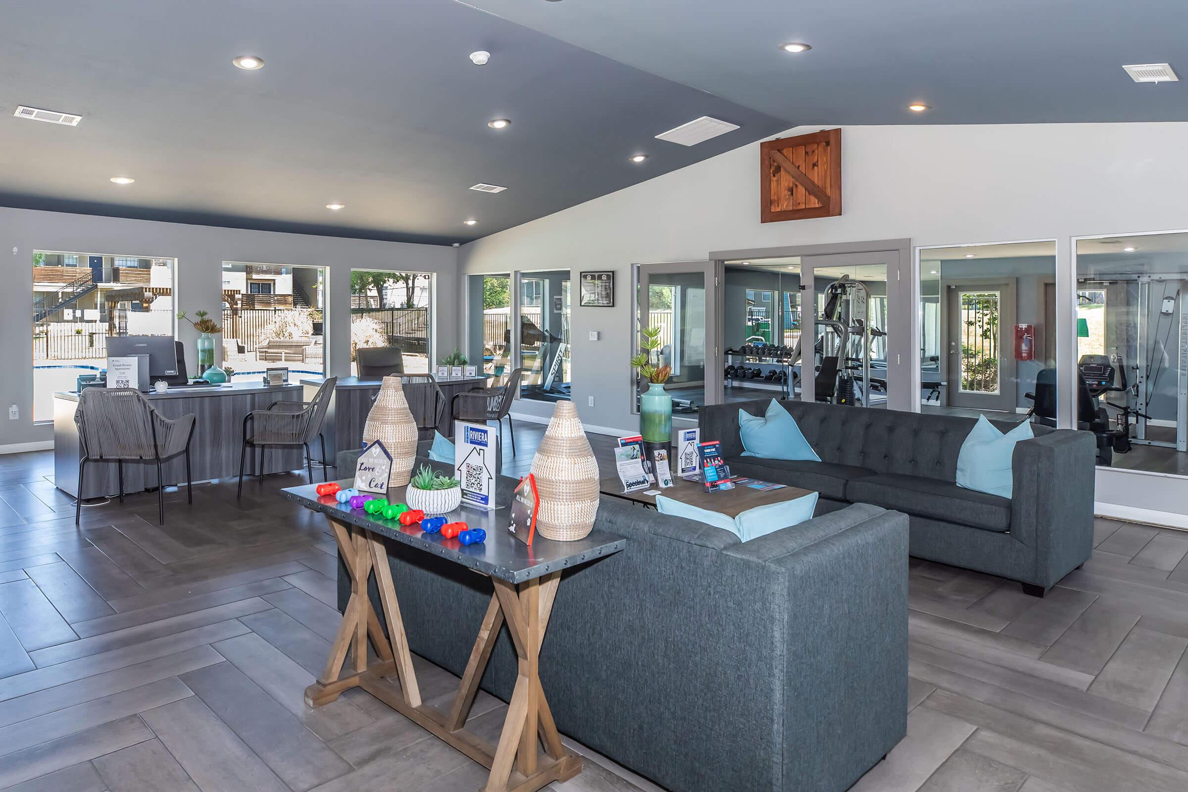 A modern community lounge featuring a gray sofa with blue cushions, a wooden table displaying informational brochures and colorful toys, and large windows letting in natural light. In the background, there's a gym area with exercise equipment and a view of a pool area outside.