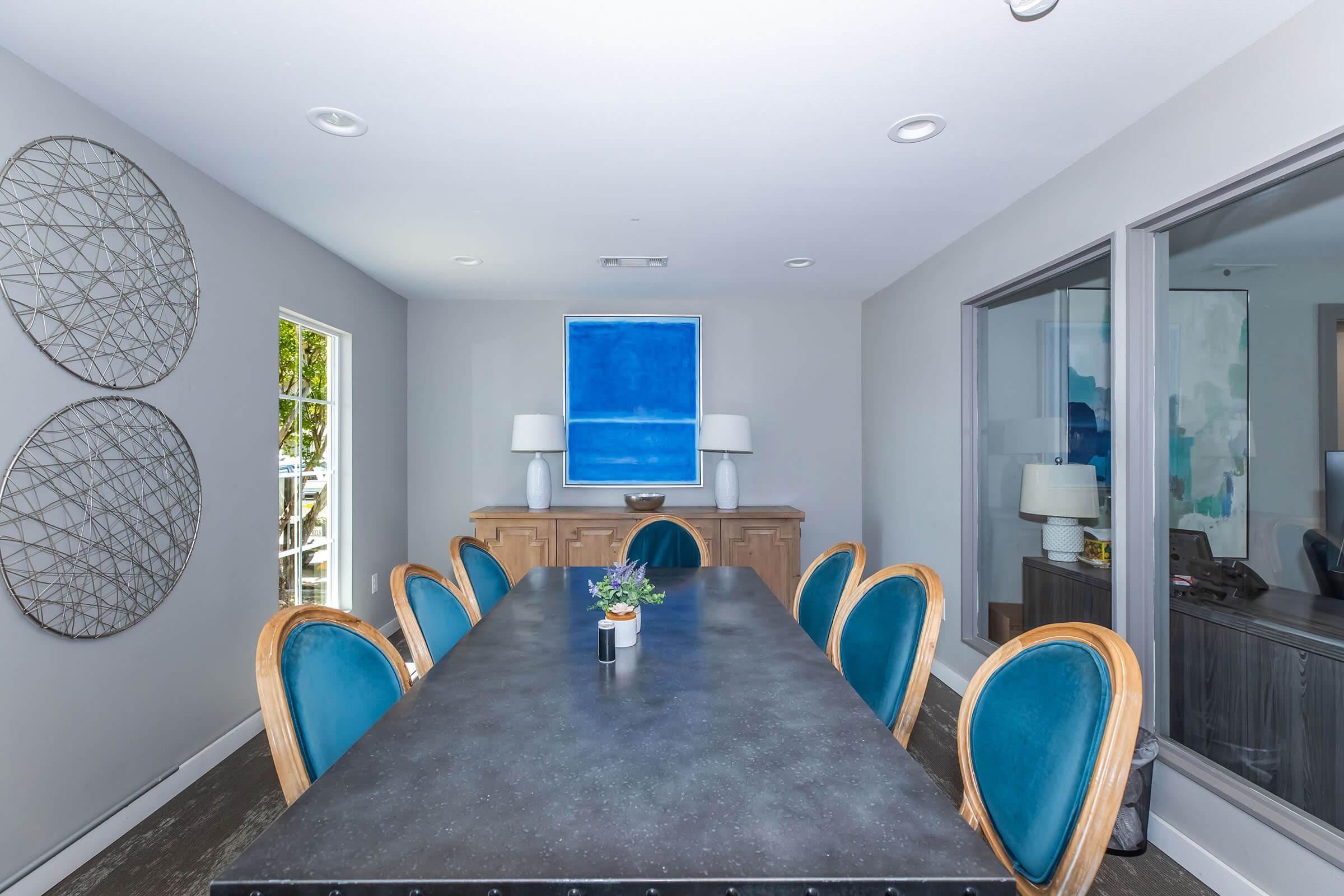 A modern dining room featuring a long table with teal upholstered chairs. Two decorative circular wall art pieces are displayed on a light gray wall. A sideboard with a blue artwork above it is visible, along with large windows that allow natural light to fill the space.