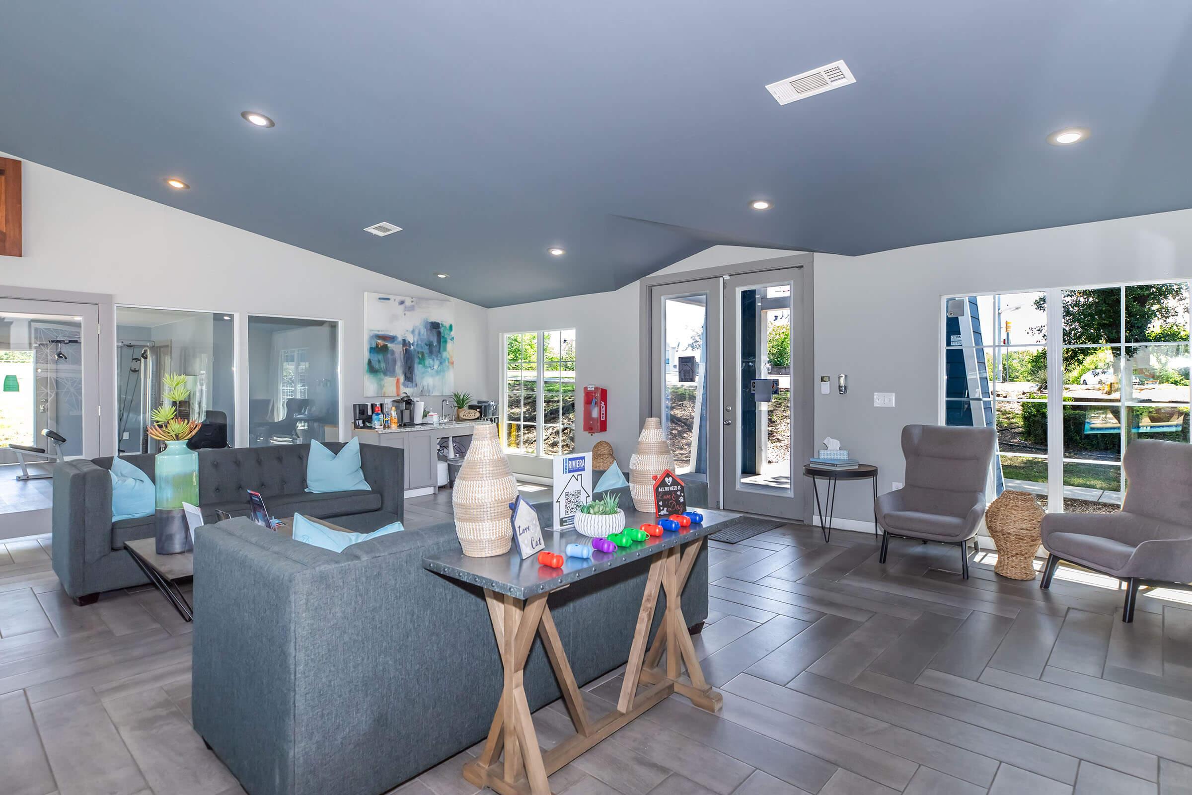 A modern living space featuring gray sofas, a coffee table with colorful objects, and large windows allowing natural light. The room has a blue ceiling, and there are two armchairs. In the background, kitchen appliances and a view of the outdoors can be seen, creating a bright and inviting atmosphere.