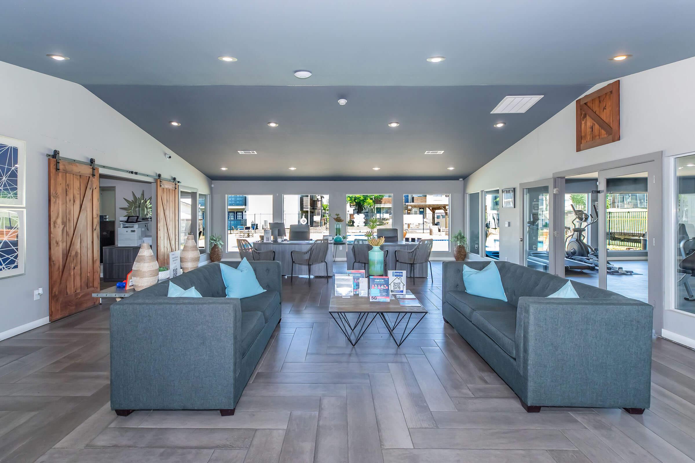 Modern lounge area featuring two gray sofas with light blue cushions facing a central coffee table. Large windows let in natural light, and sliding glass doors open to an outdoor space. The decor includes potted plants and stylish light fixtures, creating a welcoming and contemporary atmosphere.