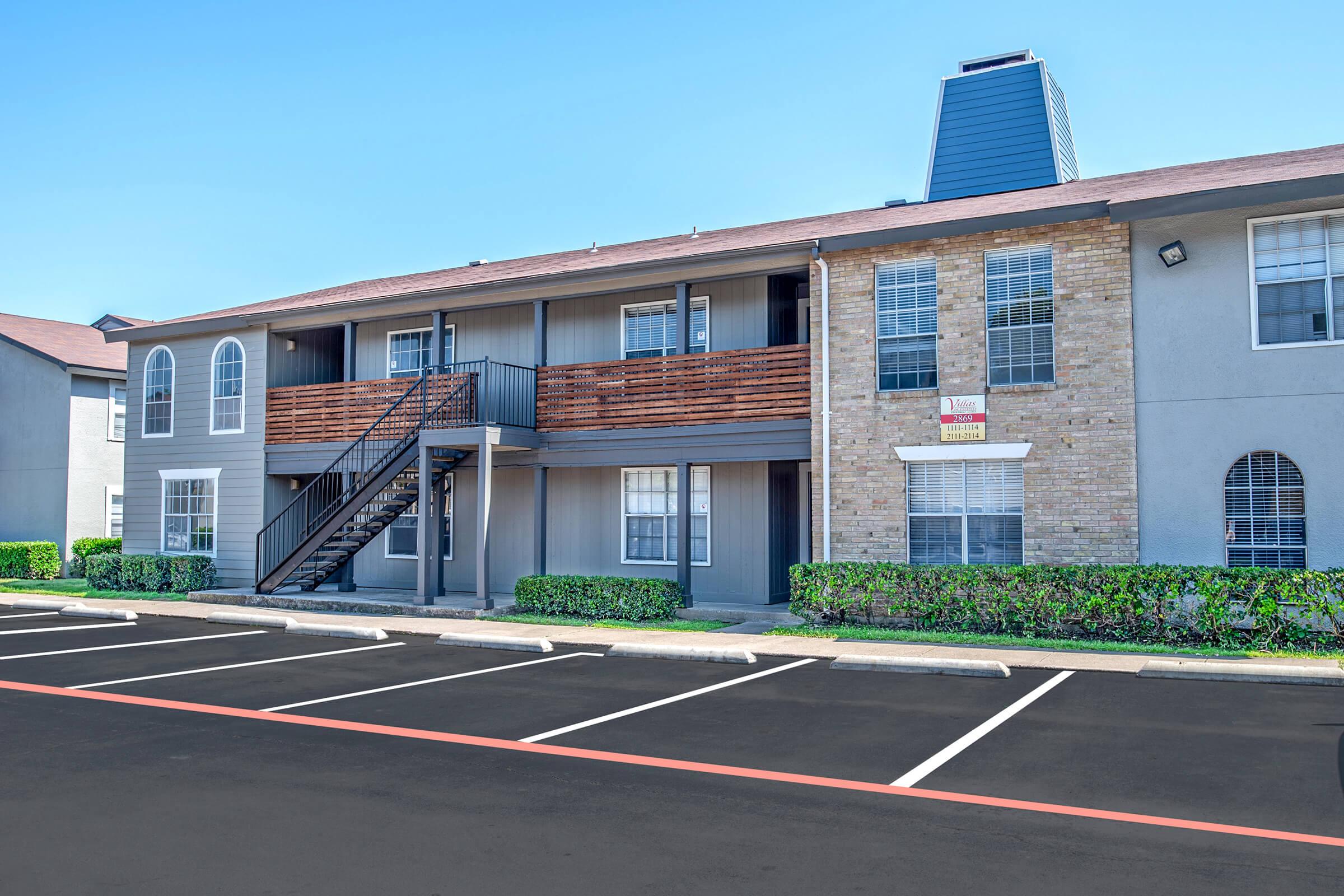 Exterior view of a two-story apartment complex featuring a brick and stucco façade, wooden balconies, and a black metal staircase. Surrounding the building are neatly trimmed shrubs and a parking lot with marked spaces. The sky is clear and blue.