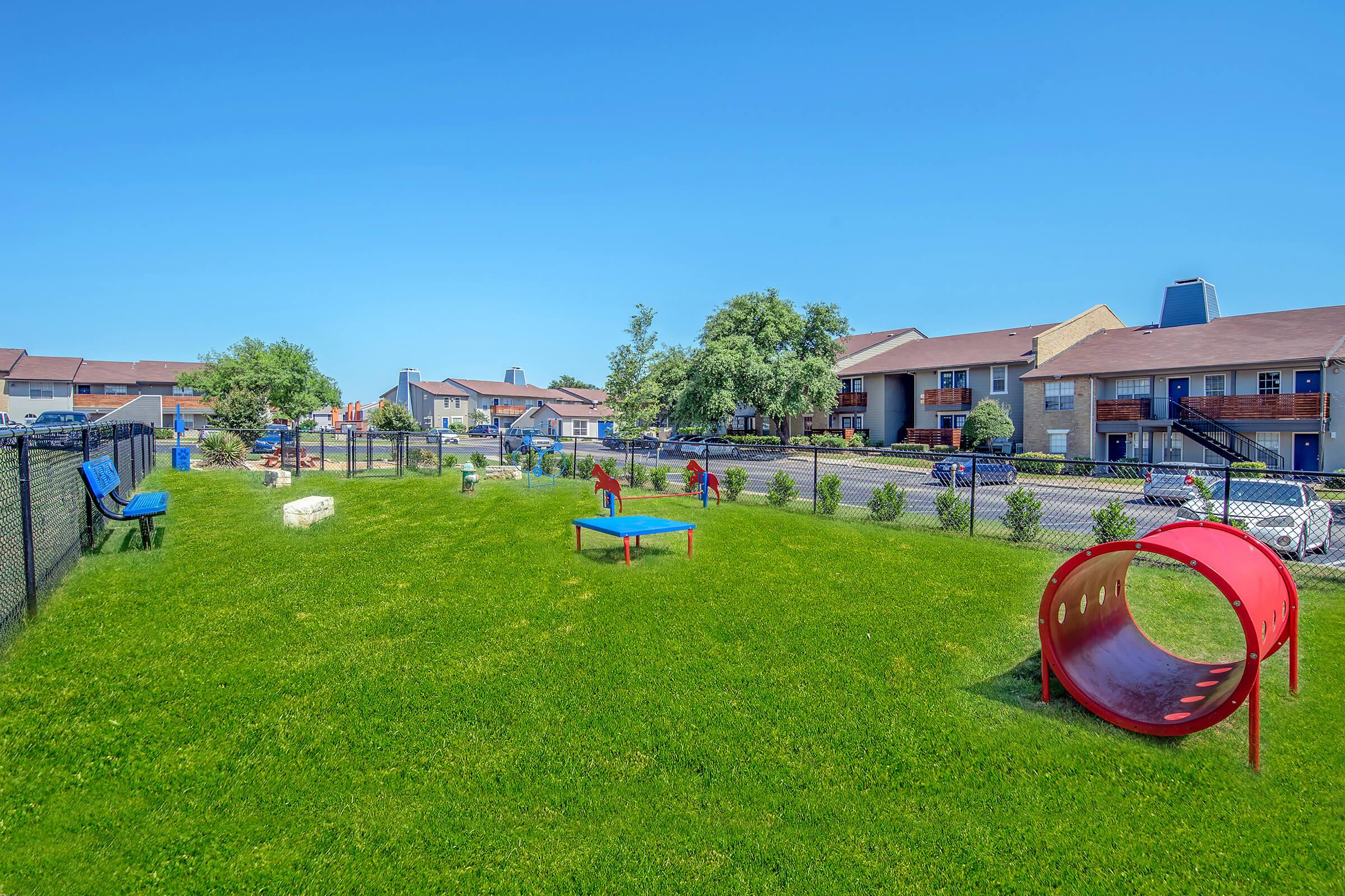 A spacious outdoor area in a residential setting featuring well-maintained green grass. The area includes playground equipment like colorful tunnels and platforms, with trees and shrubs lining the fence. Nearby, residential buildings can be seen in the background under a clear blue sky.