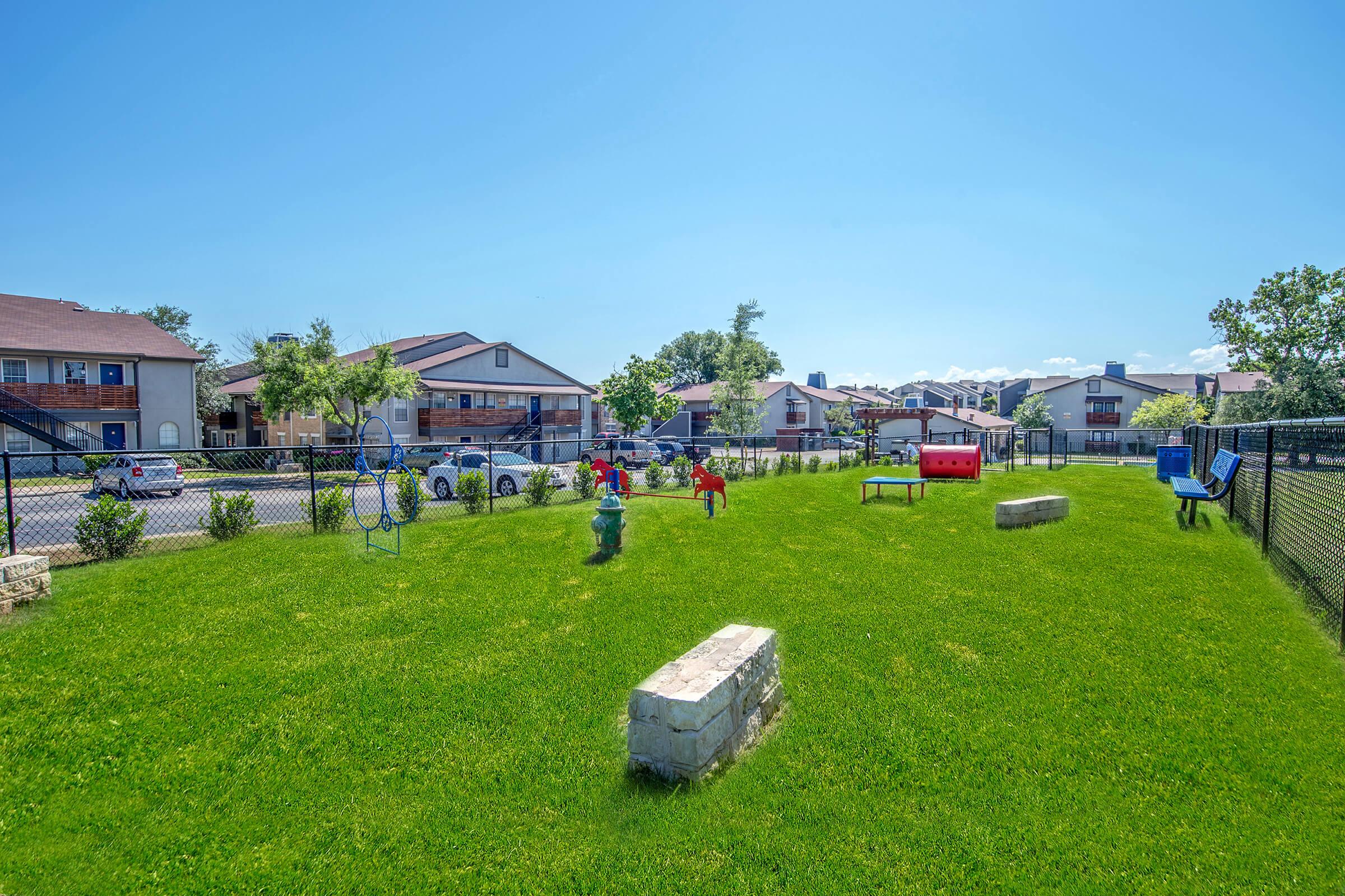 A spacious green playground with a fenced area featuring children playing. Two colorful play structures are visible, alongside benches and other equipment. In the background, residential buildings are set against a clear blue sky. The grassy area is well-maintained, providing a cheerful atmosphere for outdoor play.