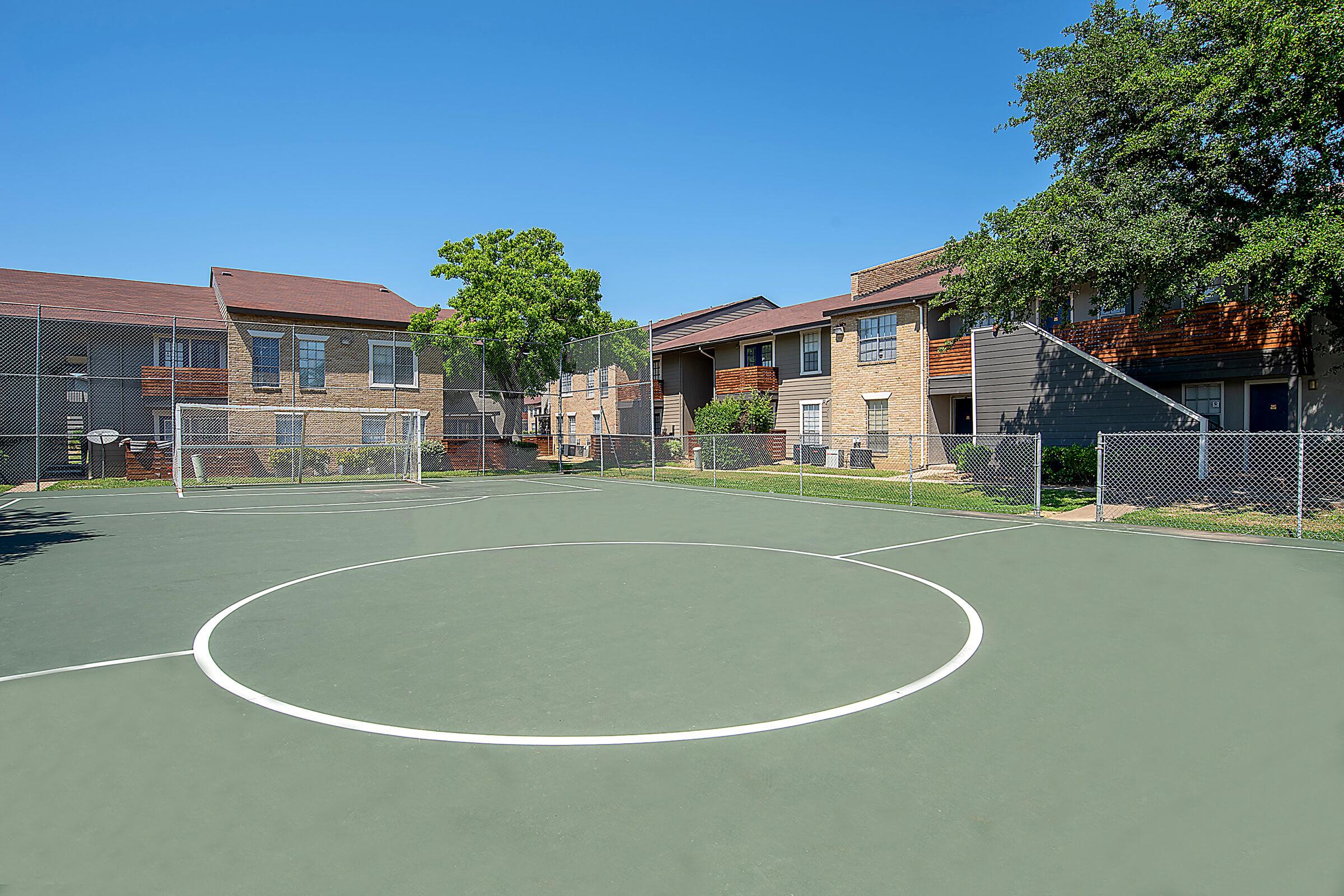 A well-lit outdoor sports court surrounded by residential buildings. The court features a circular center and is enclosed by a chain-link fence. Lush green trees provide shade nearby, and the sky is clear and blue, enhancing the recreational atmosphere of the area.