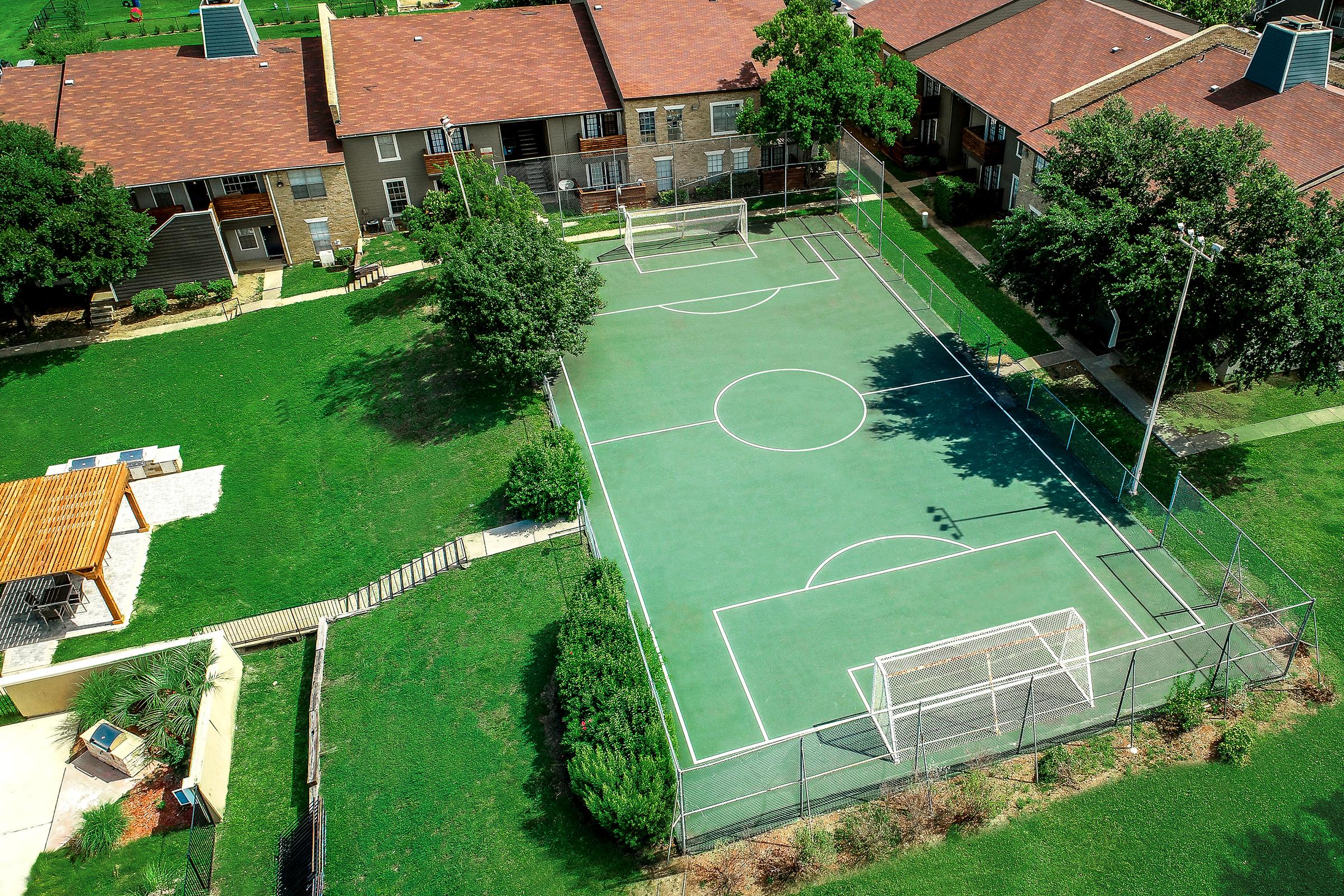 Aerial view of a green soccer field surrounded by residential buildings. The field features white goalposts at each end, a center circle, and clear boundary lines. Nearby, there are trees and a landscaped area, along with steps leading down to a lower level.