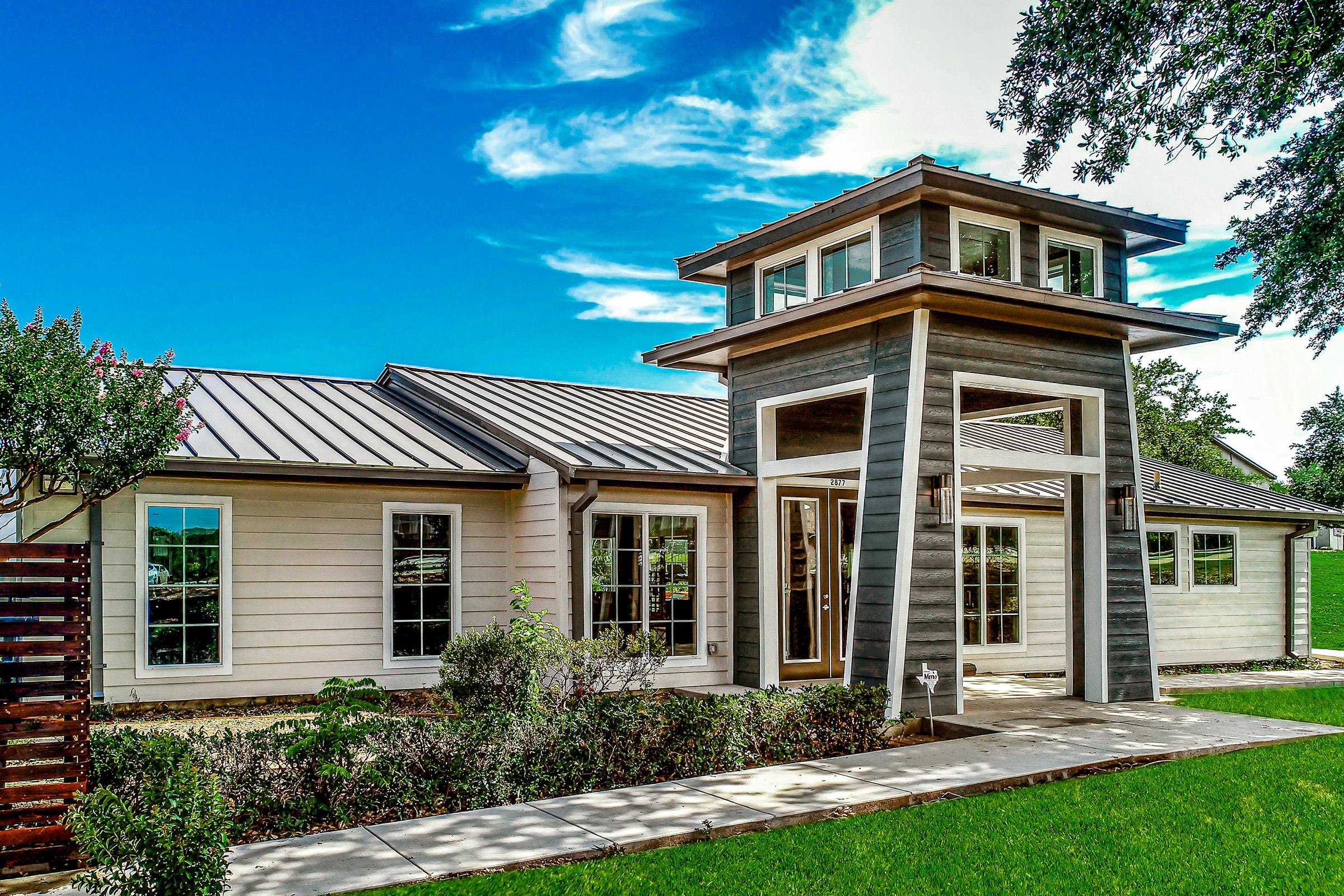 A modern two-story house featuring a unique tower-like entrance, large windows, and a combination of horizontal and vertical siding. The home is surrounded by green grass and landscaped shrubs, with a clear blue sky and scattered clouds in the background.
