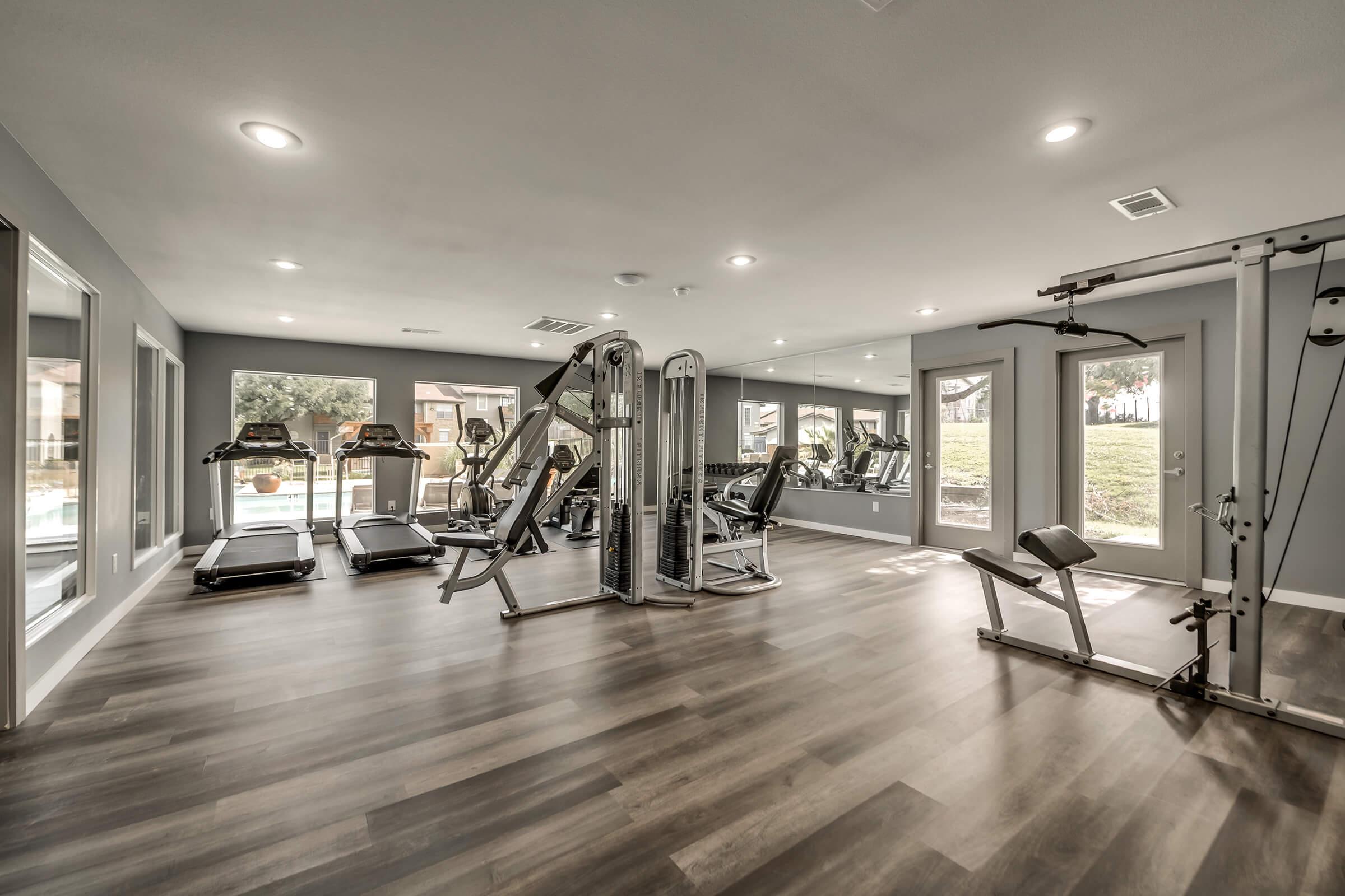 Modern home gym featuring various exercise equipment, including treadmills, weight machines, and strength training tools. The space has large windows allowing natural light, with a sleek wooden floor and a neutral color palette, creating a bright and inviting workout environment.