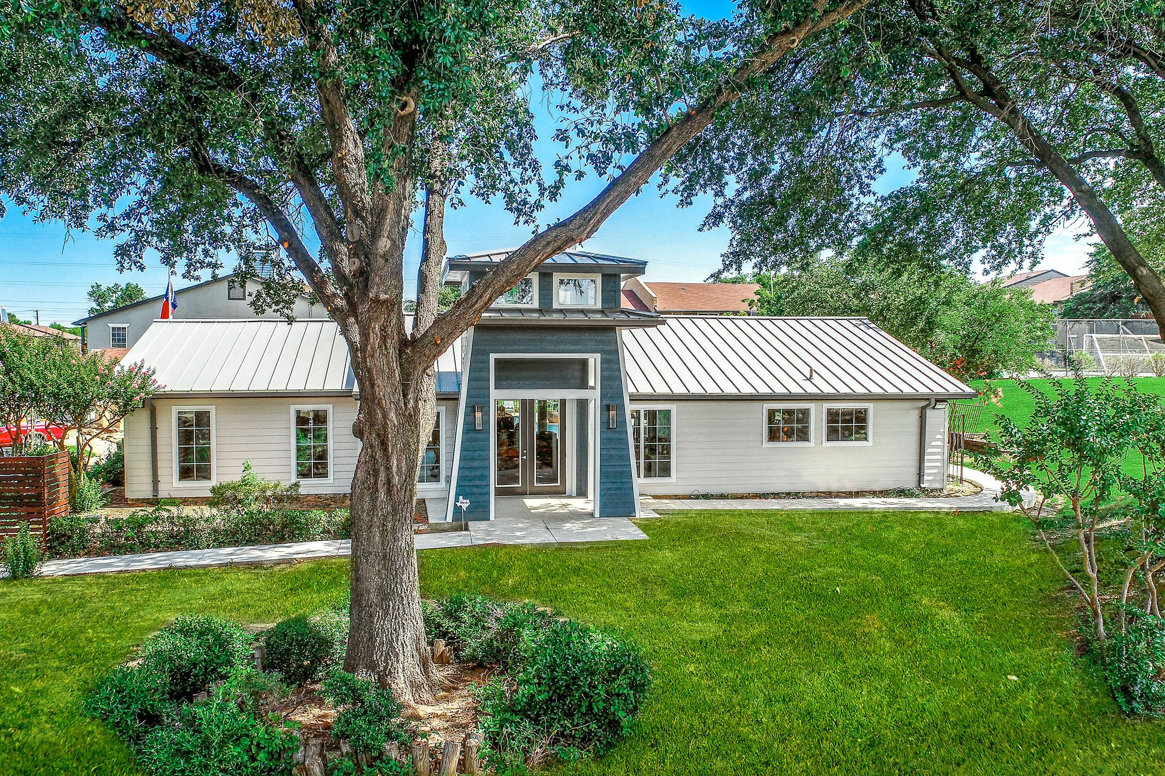 A modern single-story home with a metal roof, featuring large front windows and a welcoming entrance. Surrounded by lush green grass, manicured trees, and shrubs, the property offers a spacious yard. A driveway leads to the front of the house, enhancing its inviting appearance.