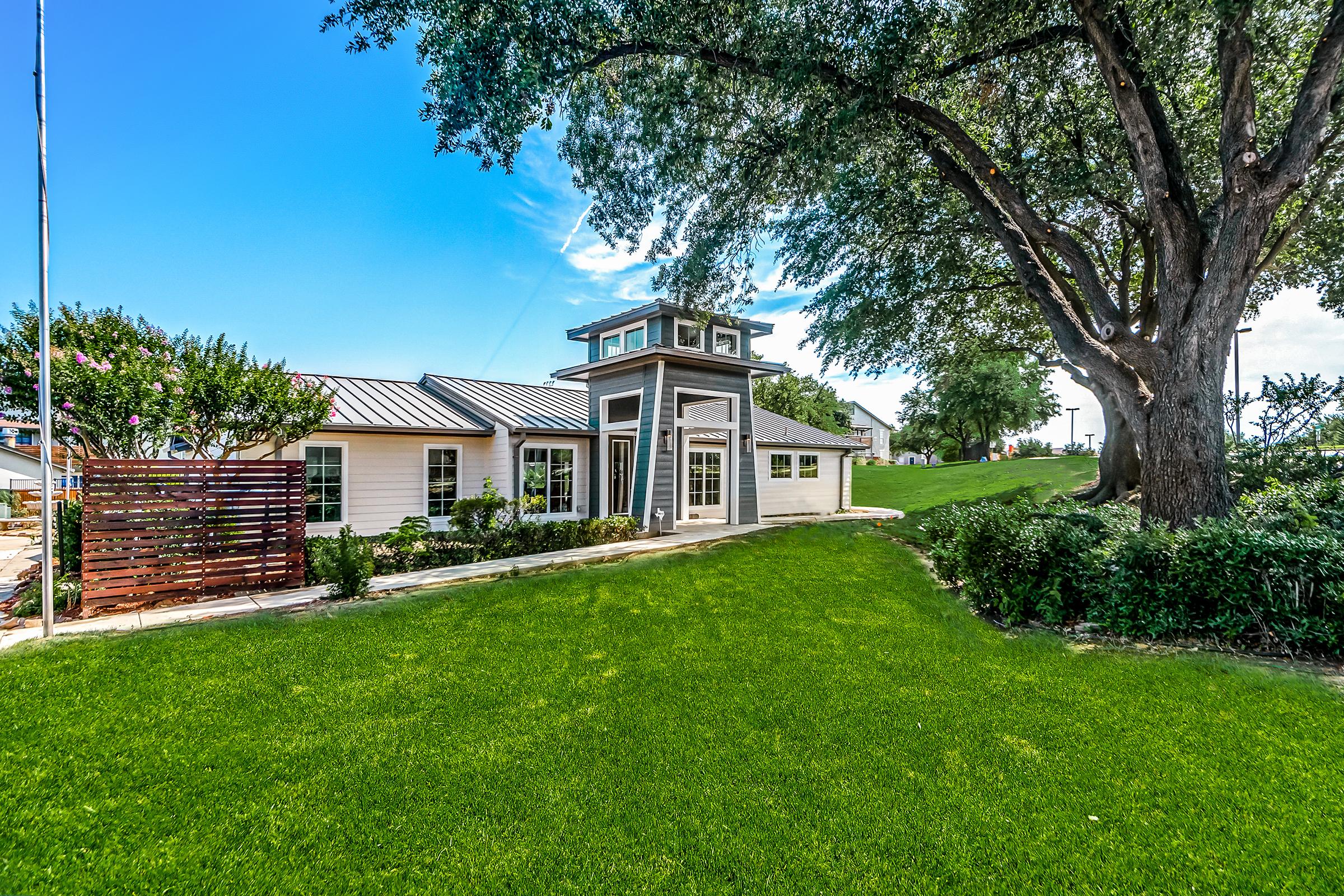 A modern house with a sleek design featuring large windows and a distinctive entrance. The property includes well-manicured green lawns and trees, creating a serene outdoor atmosphere. Bright blue sky complements the scene, enhancing the inviting aesthetic of the home.