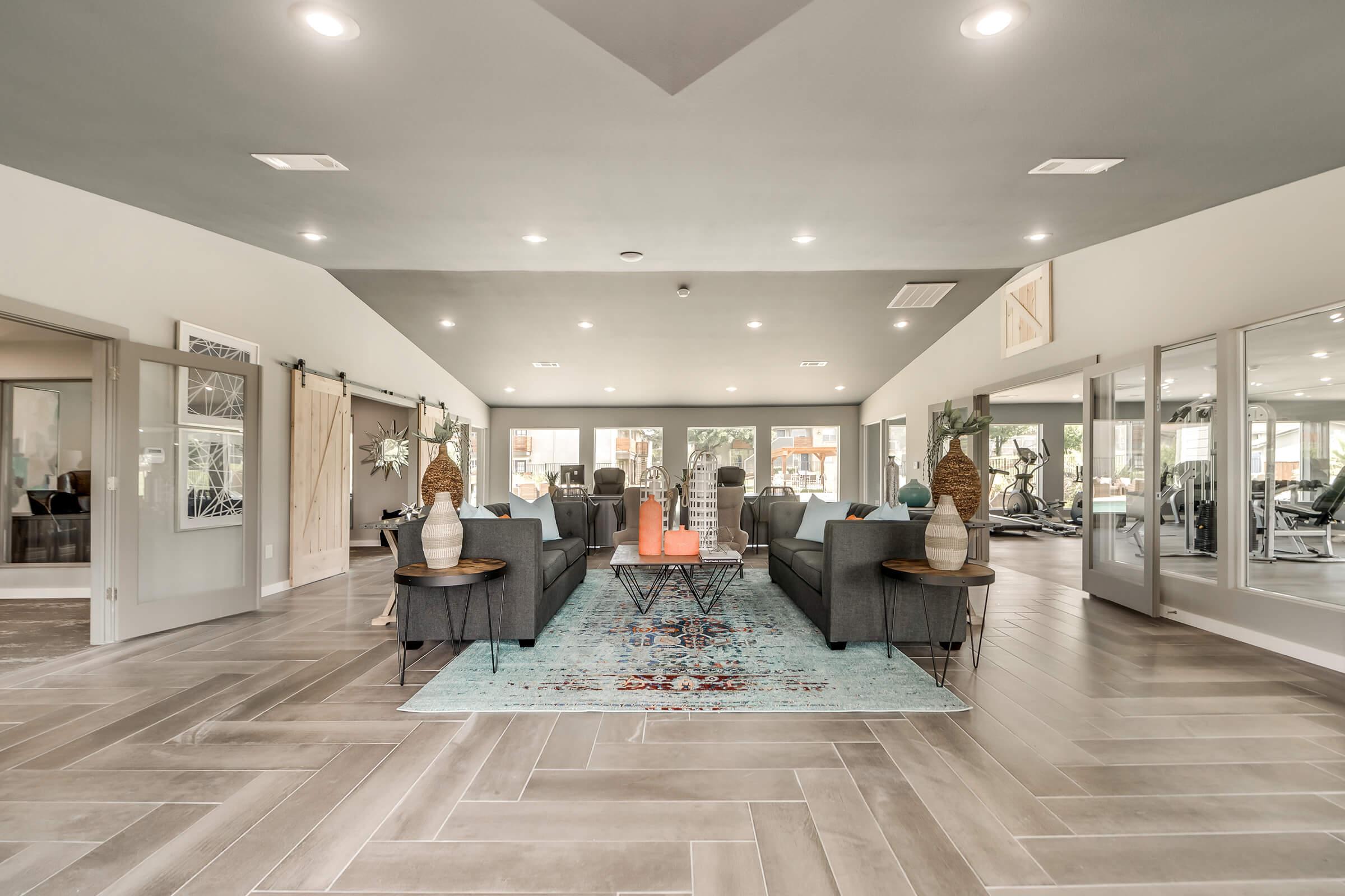 A spacious, modern lounge area featuring two gray sofas, decorative tables with vases, and a colorful rug. Large windows allow natural light to flood the room, which has a sleek, contemporary design with tiled flooring. The area is well-decorated, creating a welcoming atmosphere.