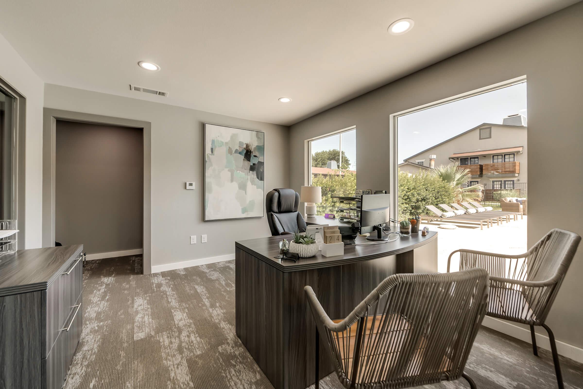 Modern office interior featuring a wooden desk with a computer and office supplies, a comfortable chair, and a large abstract painting on the wall. Windows provide natural light and a view of an outdoor pool area. The walls are painted in neutral tones, enhancing a bright and inviting atmosphere.