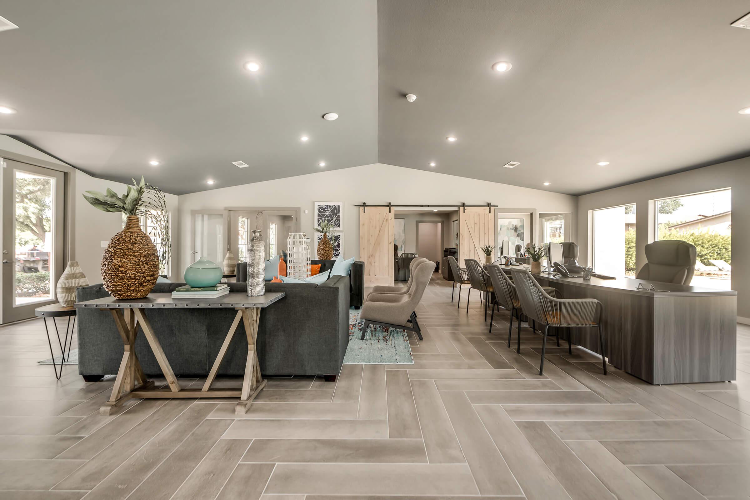 Interior view of a modern lounge area featuring a variety of seating options, a wooden reception desk, and decorative elements like plants and vases. Large windows allow natural light to fill the space, complemented by stylish flooring and contemporary design accents.