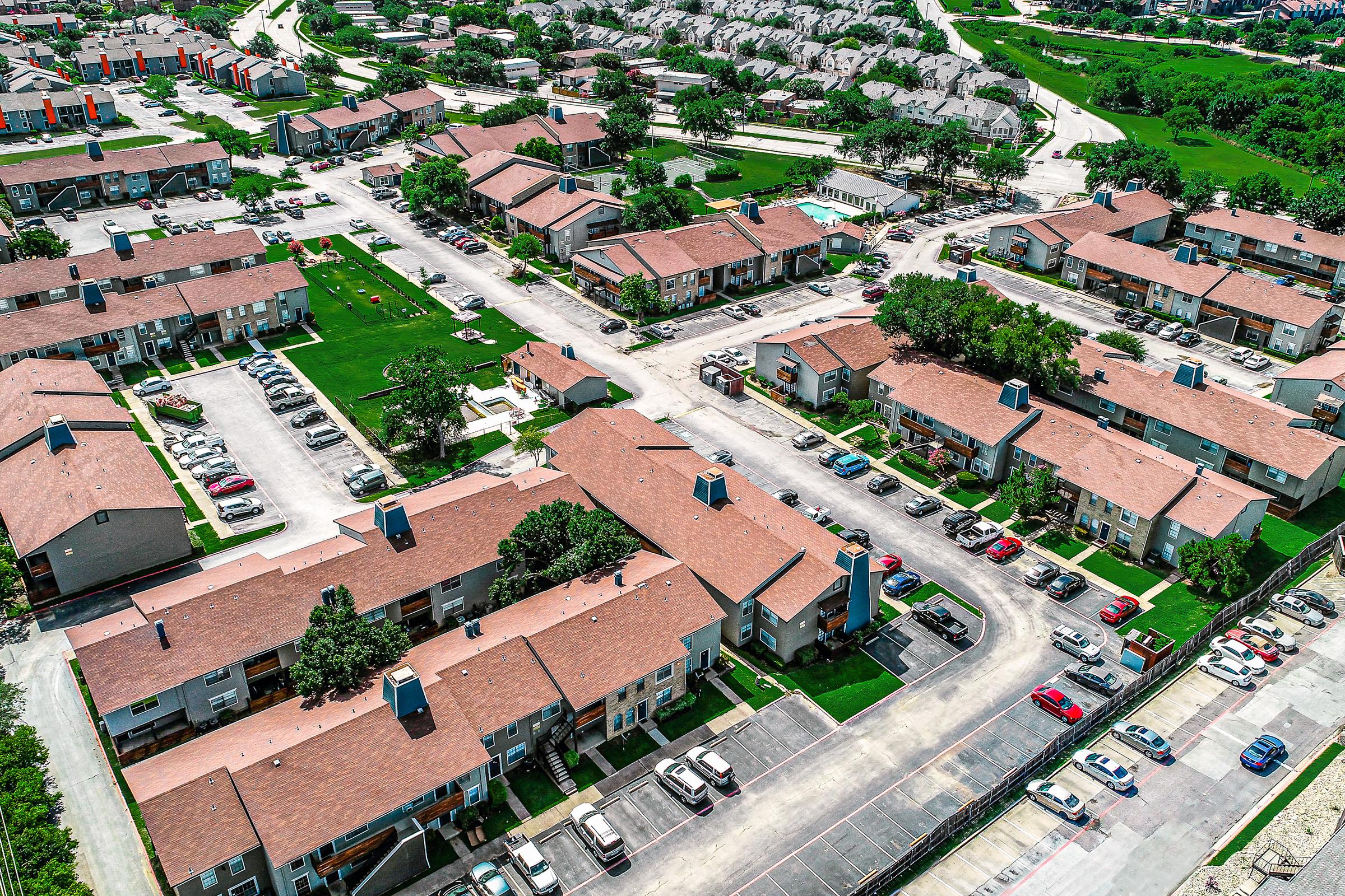 Aerial view of a residential community featuring multiple apartment buildings with brown roofs, grassy areas, and parked cars. The layout shows several streets, green spaces, and amenities like a swimming pool in the background, set against a suburban landscape.