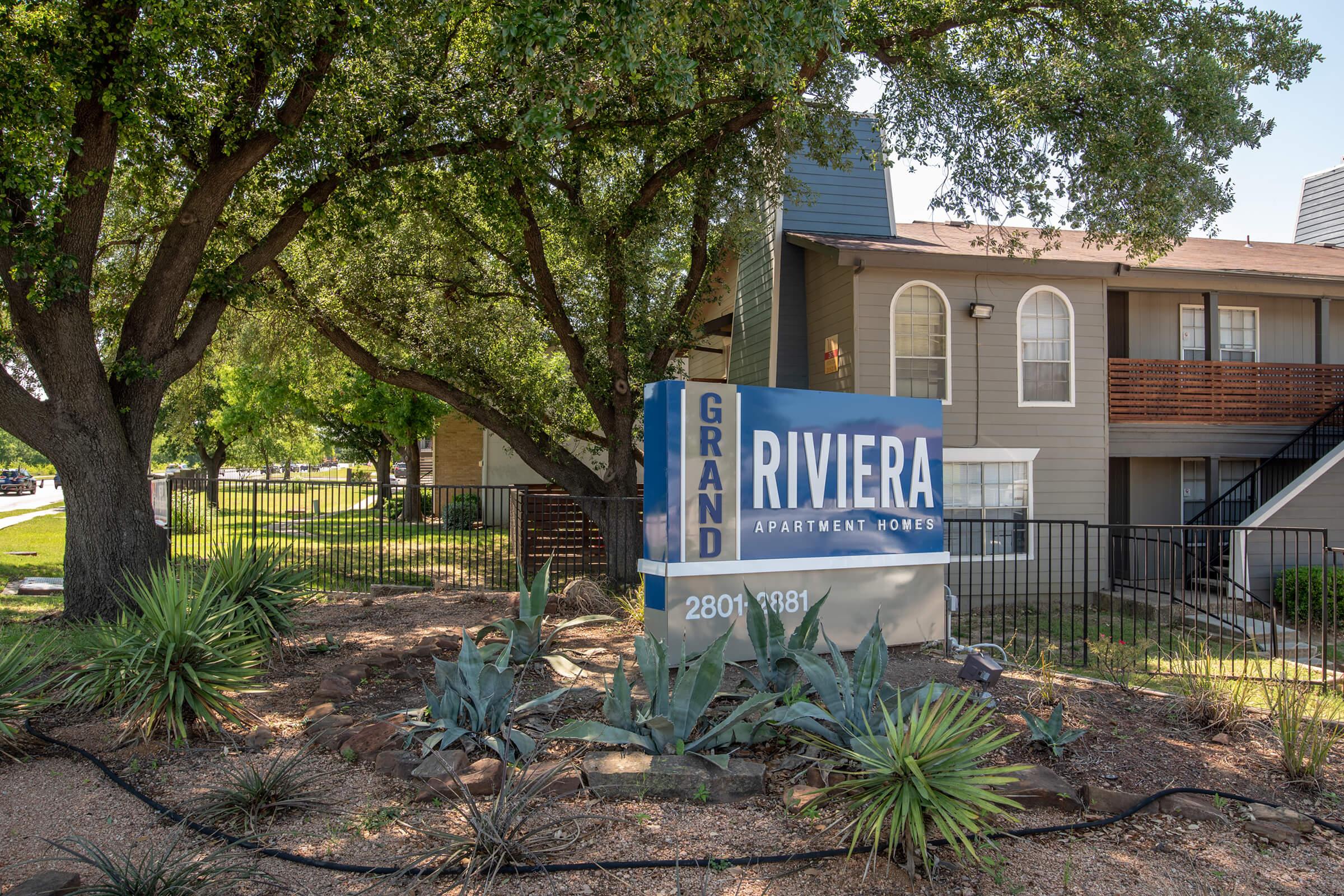 Sign for Grand Riviera Apartment Homes, featuring blue and white colors, set in a landscaped area with greenery. In the background, there are residential buildings. The sign displays the address "2800" prominently.