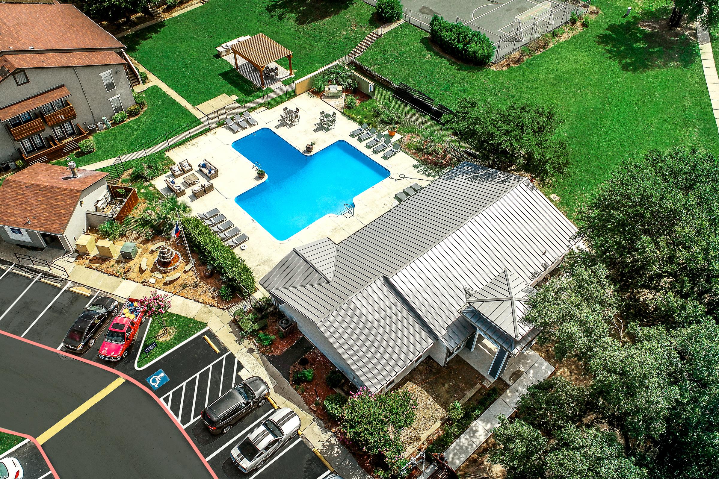 Aerial view of a community pool area featuring a rectangular blue swimming pool surrounded by lounge chairs. Nearby, there's a clubhouse with a metal roof, green lawns, and a well-maintained landscape. Parking spaces are visible in the foreground and additional amenities, like a barbecue area, are nearby.