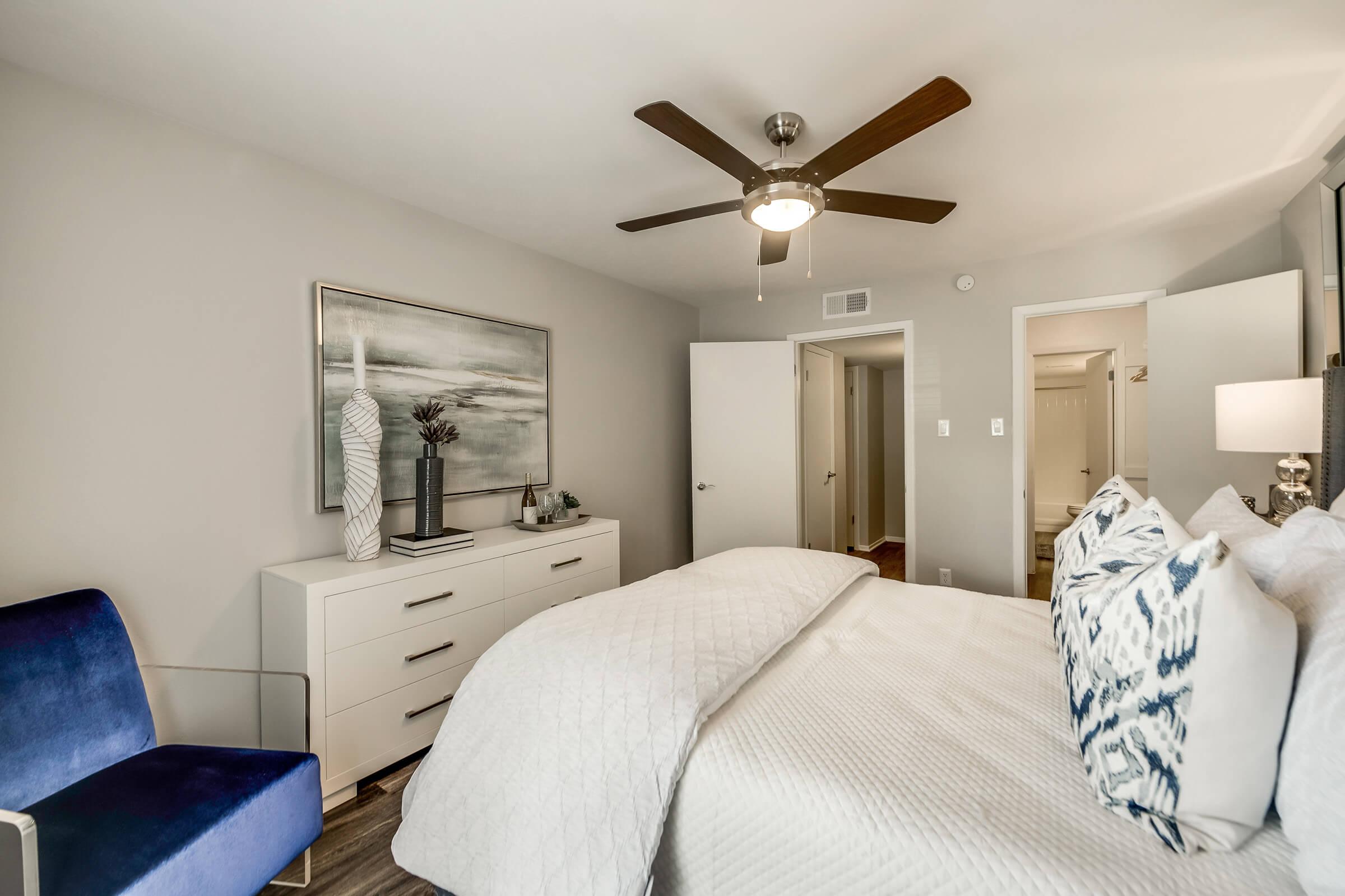 A modern bedroom featuring a neatly made bed with decorative pillows, a stylish blue chair, and a white dresser. There's a ceiling fan above, light gray walls, and artwork above the dresser. A door leads to a bathroom, and natural light fills the room, creating a cozy atmosphere.