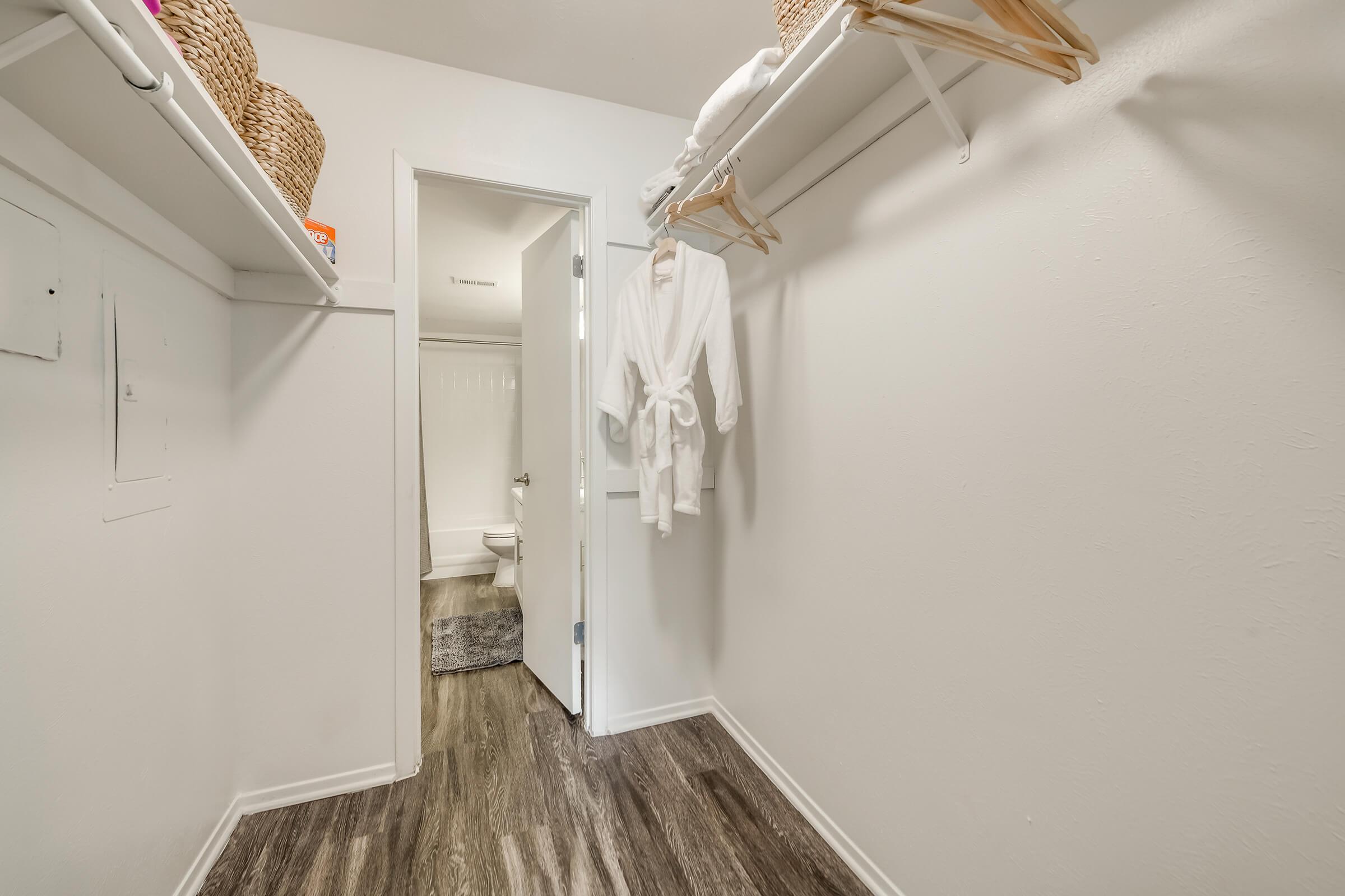 A neatly organized walk-in closet featuring white walls, wooden flooring, and shelves. Two woven baskets are positioned on the upper shelves, while a white bathrobe hangs on a wooden hanger. A door at the end of the closet leads to a bathroom, partially visible.