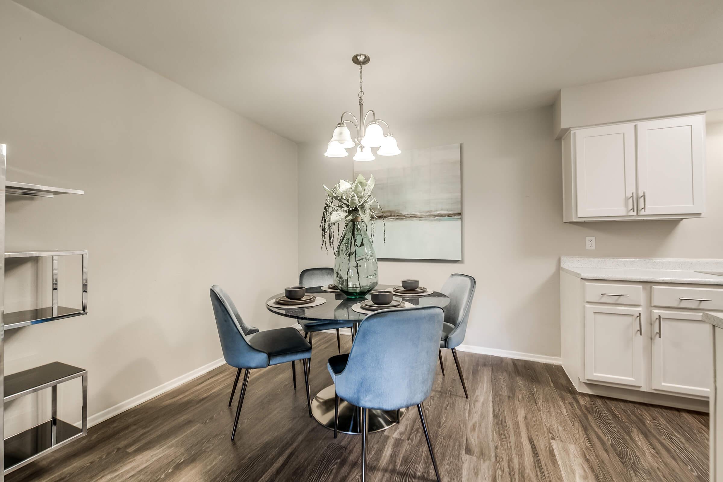 A modern dining area featuring a round glass table with four blue upholstered chairs. A large vase with greenery serves as the centerpiece, while a painting is displayed on the wall. The kitchen area has white cabinets and a sleek countertop, with a stylish light fixture overhead illuminating the space.