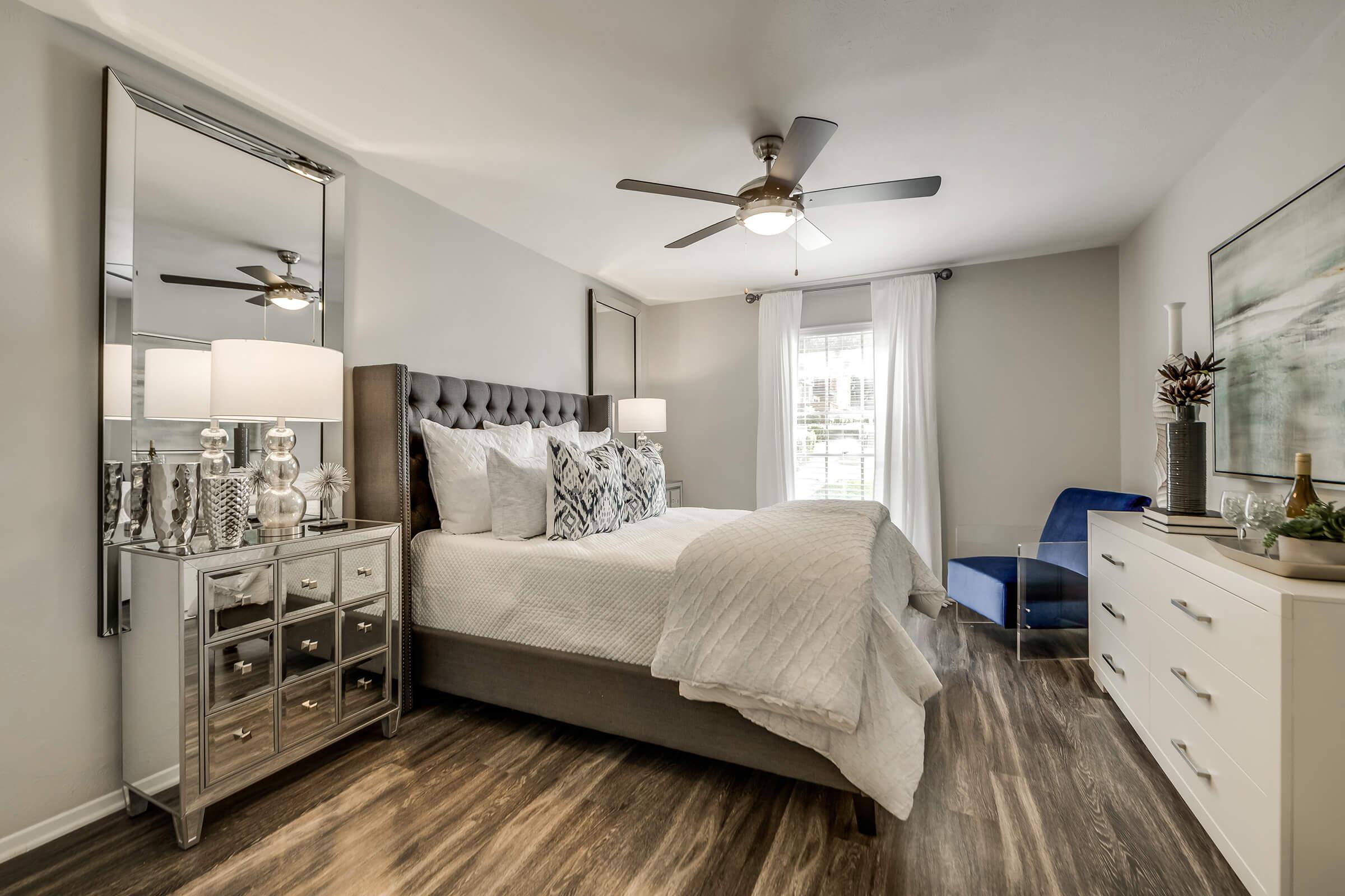 A cozy bedroom featuring a tufted gray headboard, white bedding, and decorative pillows. There are mirrored nightstands with lamps on either side of the bed, a blue accent chair in the corner, and a modern dresser. Natural light filters in through a window with white curtains, enhancing the inviting atmosphere.