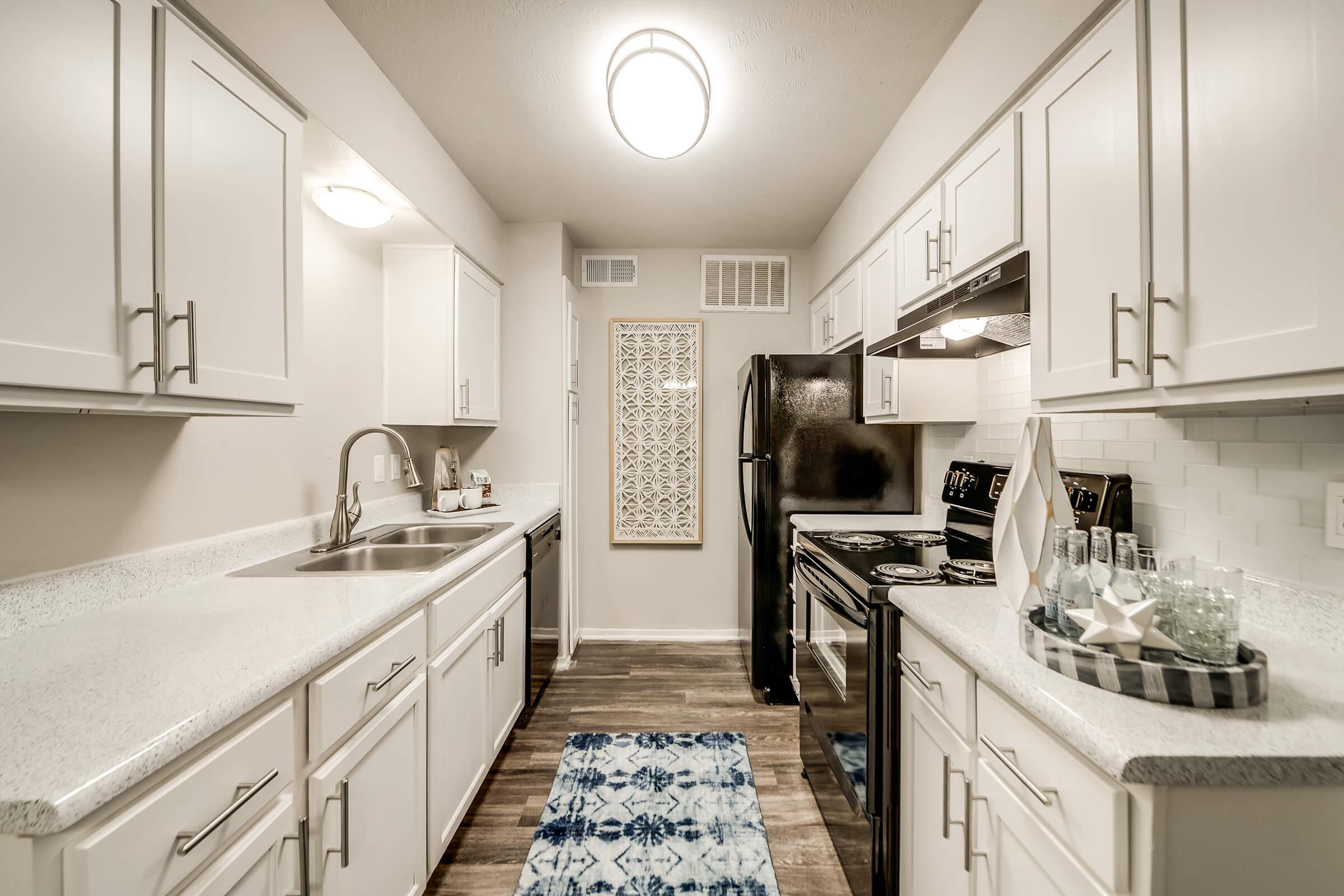 A modern kitchen featuring white cabinets, a black refrigerator, and an oven. The countertops are light-colored with a subtle texture, and a decorative wall panel is displayed. A blue and white patterned rug lies on the wooden floor, complementing the overall clean and contemporary design.