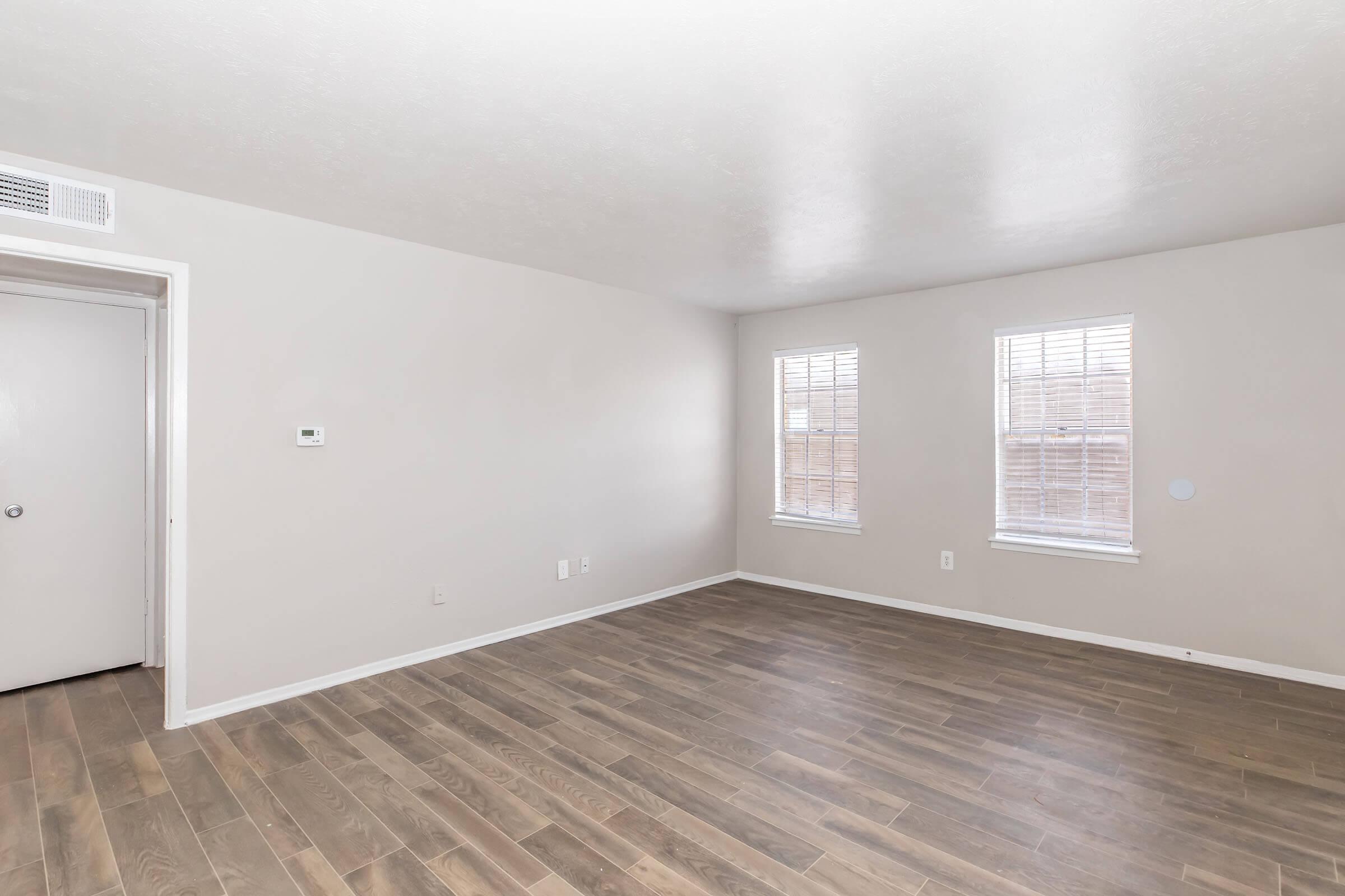 Empty room with light-colored walls and wooden flooring. Two windows with blinds allow natural light in. A door leads out of the room. The space is uncluttered and appears freshly painted, providing a neutral backdrop suitable for various decor styles.