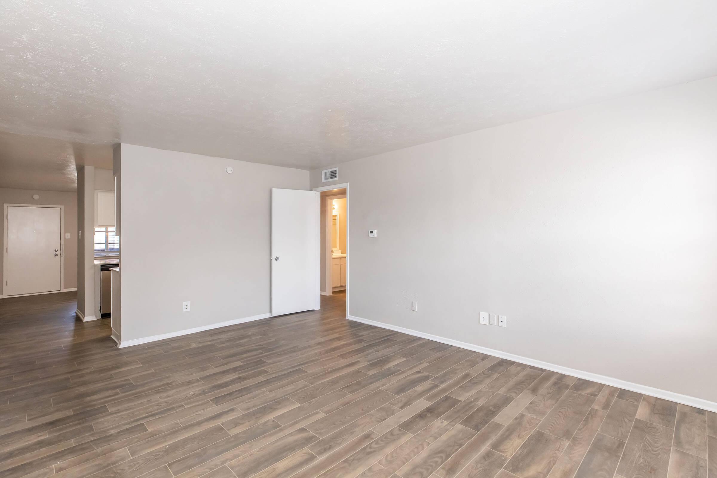 A spacious, empty room with light-colored walls and wooden flooring. The room features a closed white door on the left and an open doorway leading to a bathroom or kitchen area in the background. Natural light brightens the space, creating a warm and inviting atmosphere.