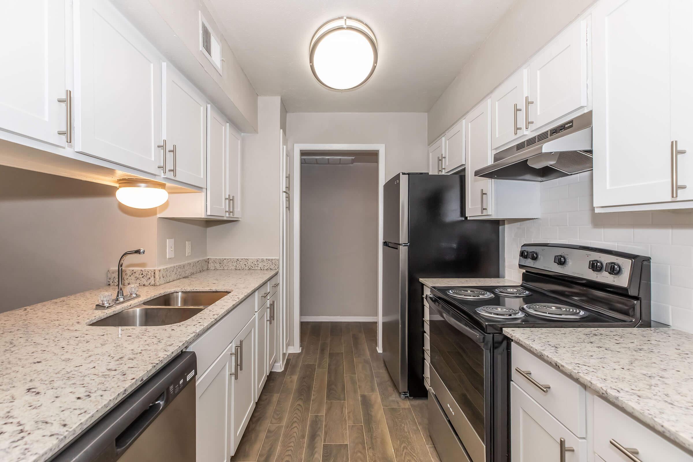 Bright modern kitchen featuring white cabinets, a double sink, and granite countertops. Equipped with a black refrigerator, a stainless steel stove, and an oven. The flooring is wood-like laminate, and there is a light fixture overhead, creating a clean and inviting atmosphere.