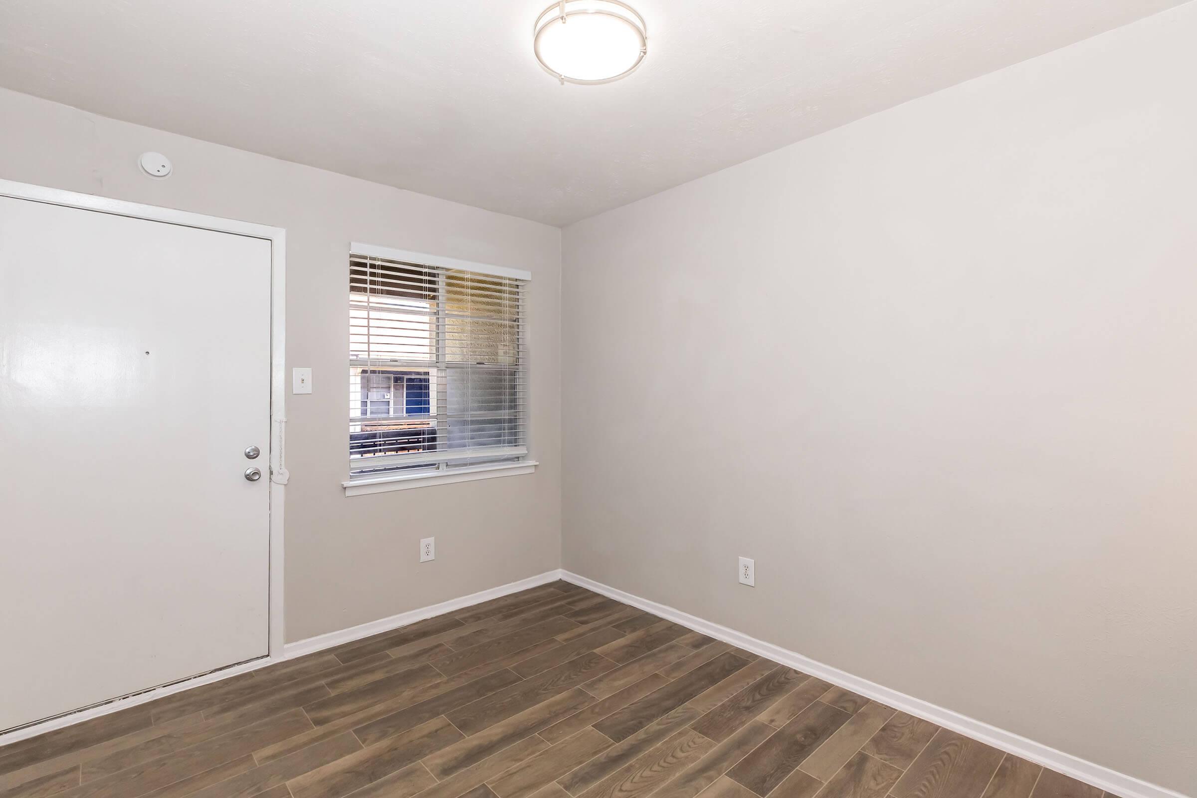 A small, empty room with light-colored walls and hardwood flooring. There is a white door on the left side and a window with blinds, allowing natural light to enter. A ceiling light fixture hangs in the center of the room, enhancing the bright and airy feel.