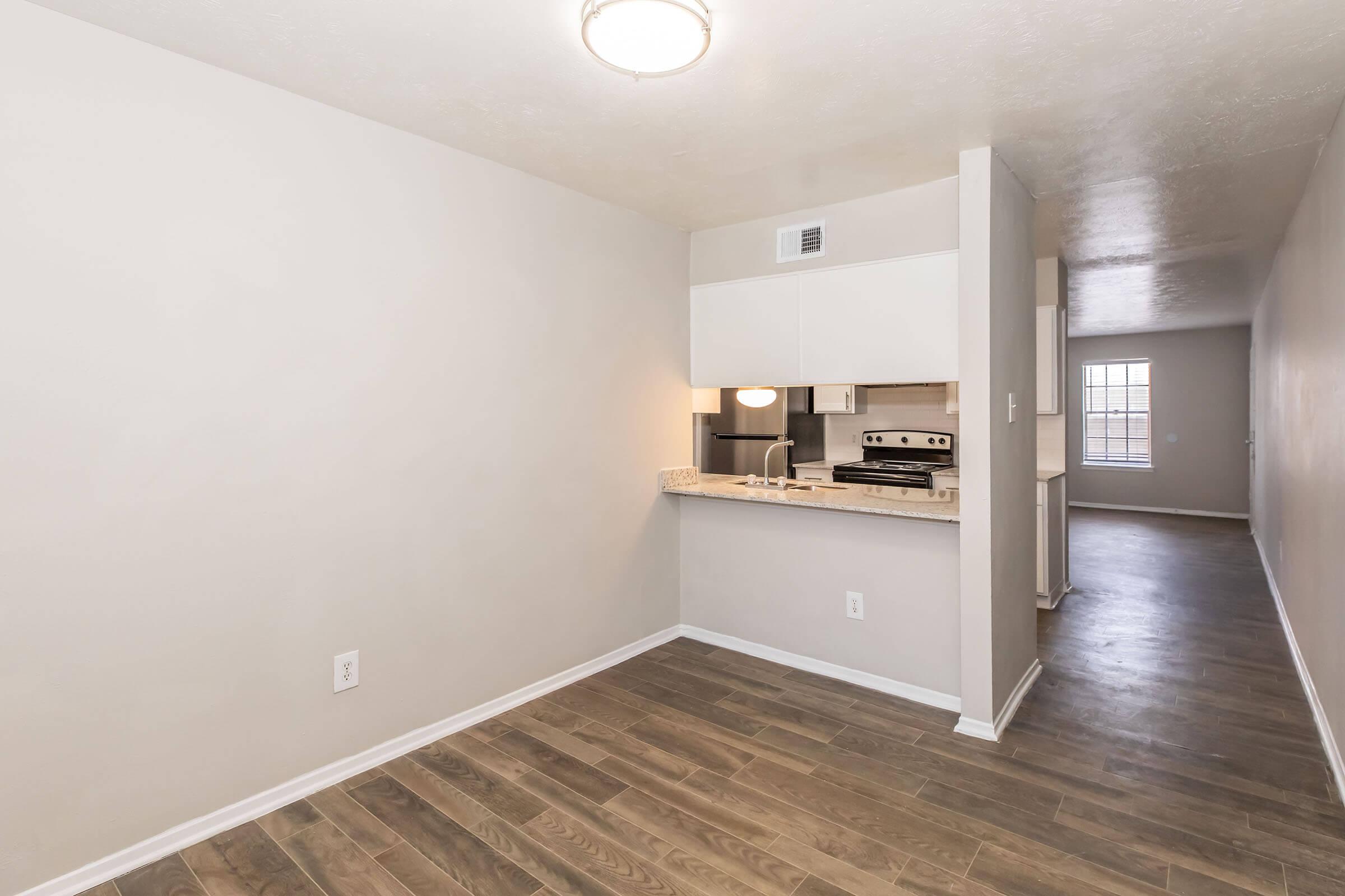 A polished interior view of an apartment showcasing a kitchen area with granite countertops, stainless steel appliances, and a light fixture. The space has light-colored walls and hardwood floors, with an open layout leading to another room visible in the background.
