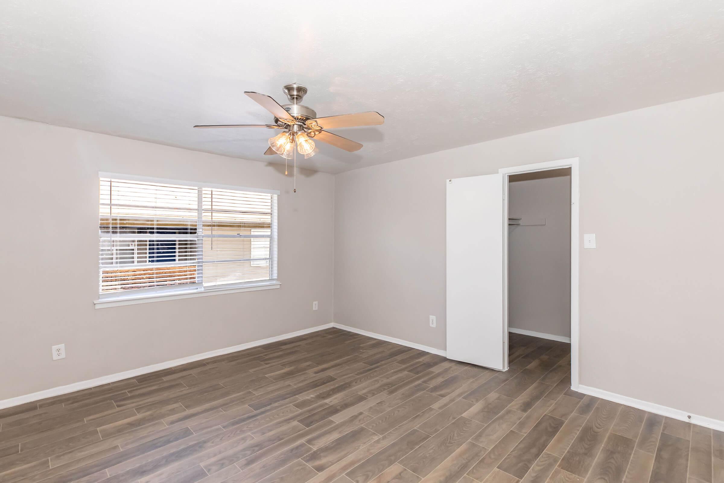 Empty room with light-colored walls and a ceiling fan. A large window with blinds lets in natural light. There is a closet with a white door on one side of the room, and the flooring is made of wooden tiles. The room appears spacious and well-lit.