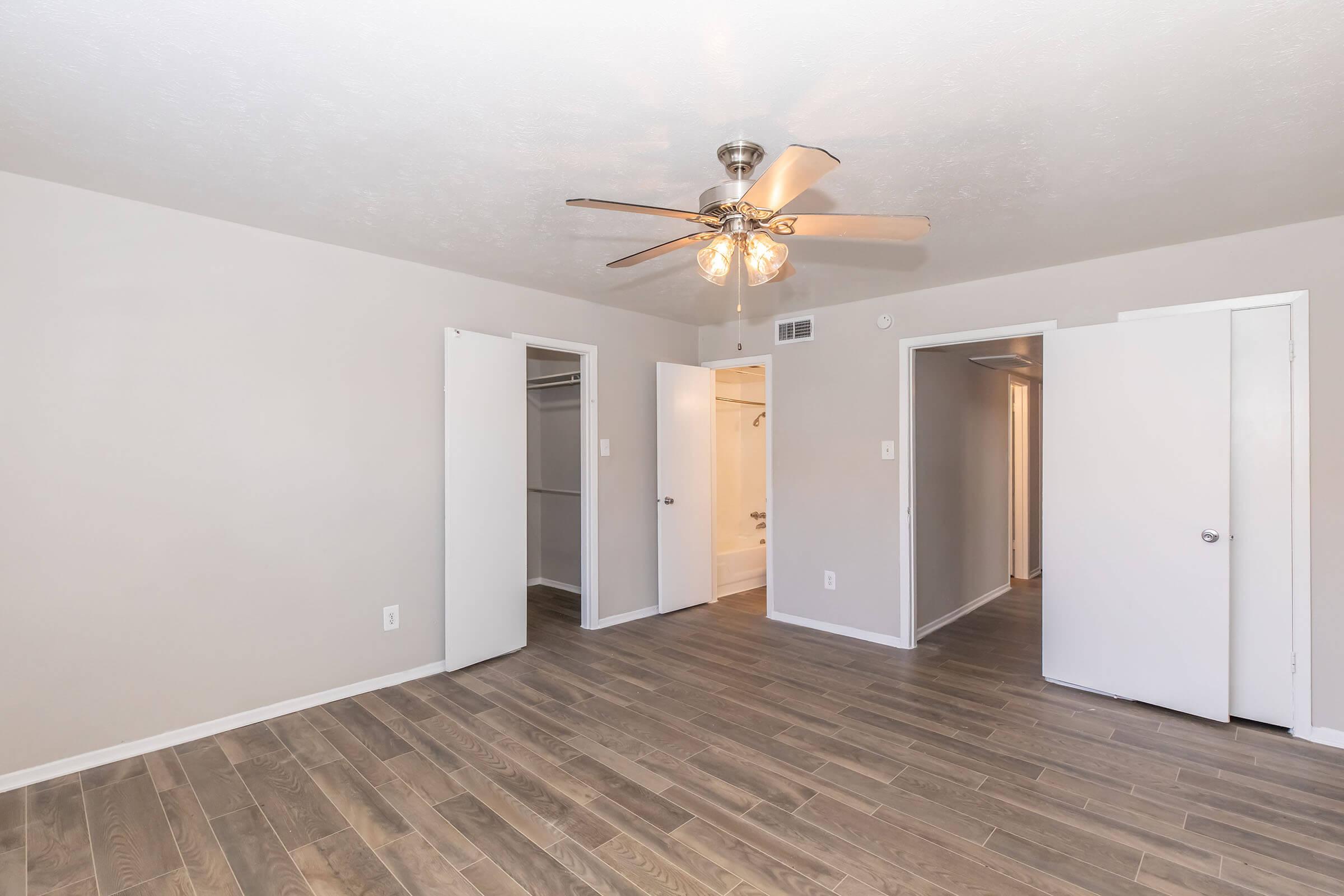 An interior room featuring light-colored walls and a wooden floor. There is a ceiling fan with a light fixture and several doorways leading to other areas. The room is well-lit and spacious, creating a comfortable atmosphere.