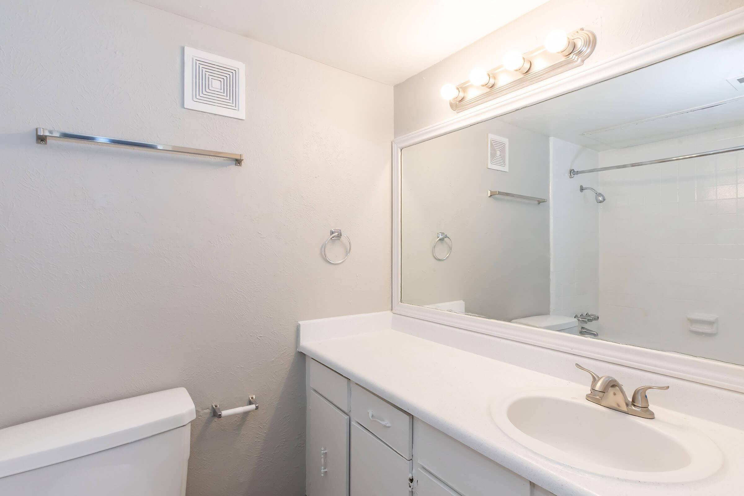 A clean, well-lit bathroom featuring a white countertop with a single sink, a large wall mirror, a towel rack, and a shower area. The walls are painted in a light color, and the fixtures are modern and simple. The overall design is minimalistic and functional.