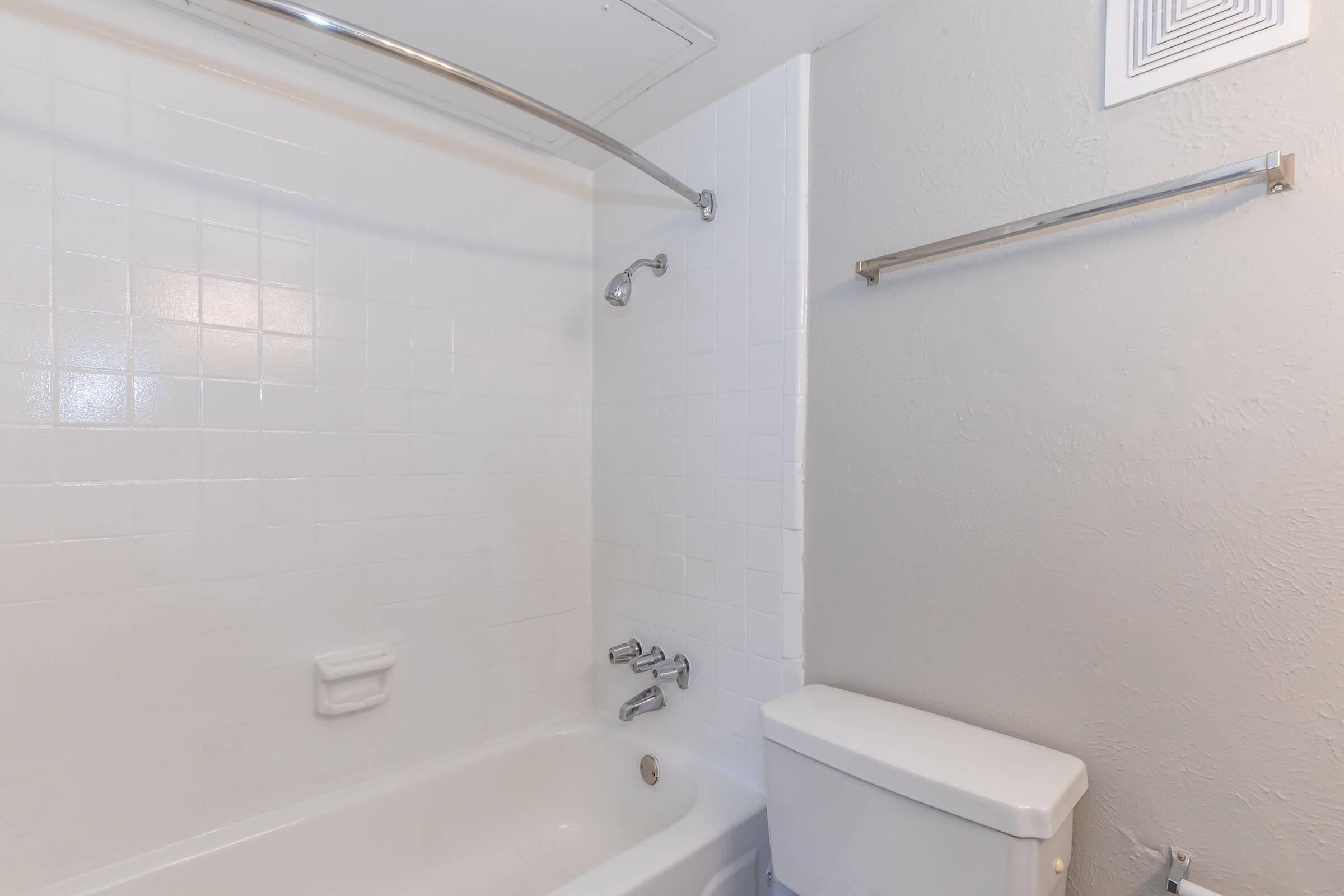 A clean and simple bathroom with a white tiled bathtub, showerhead, and a toilet. There is a shower curtain rod installed above the tub, and a wall-mounted towel rack. The walls are painted in a light color, creating a bright and spacious feel.