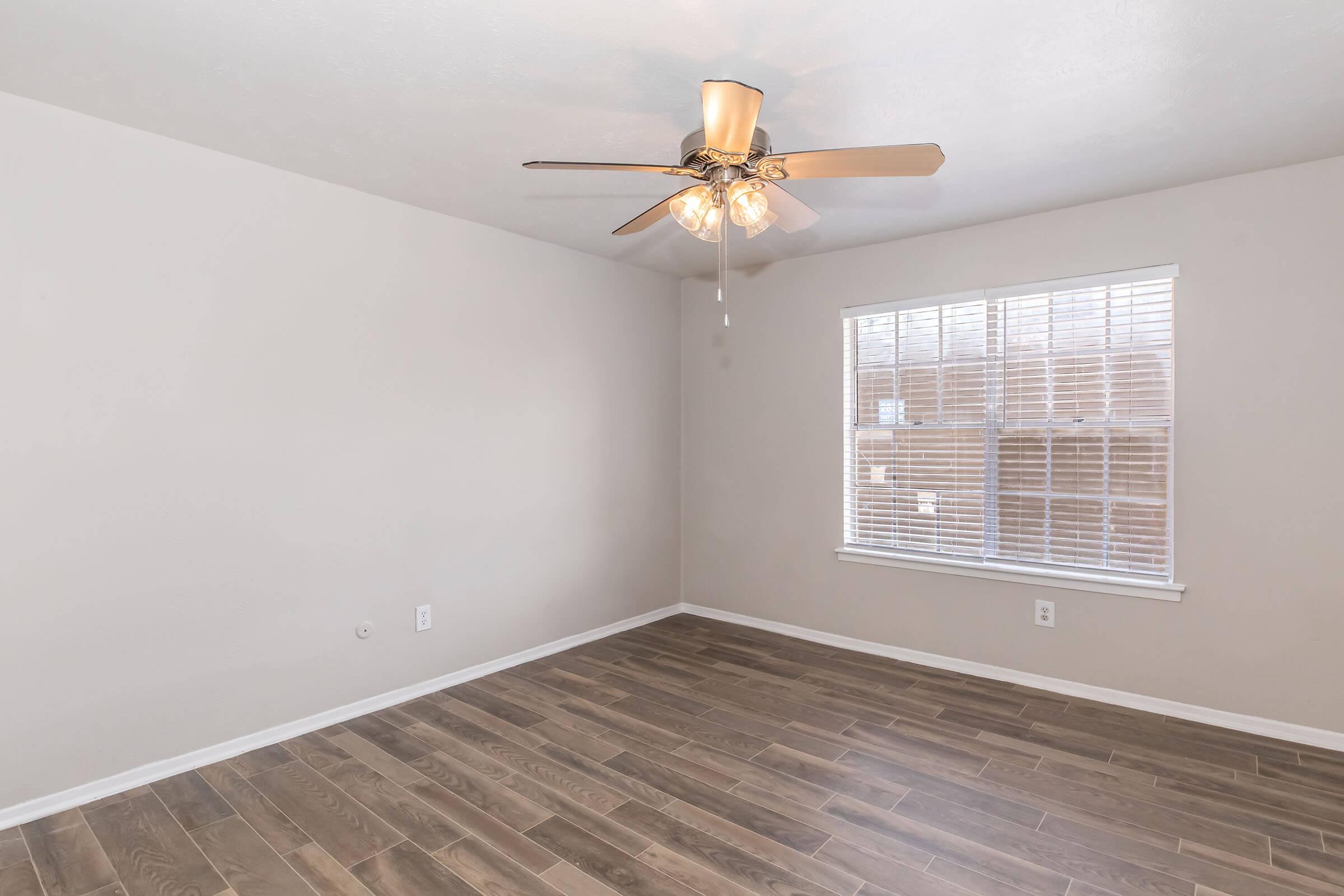 A spacious, light-filled room with pale beige walls and wooden flooring. A ceiling fan with multiple lights is mounted in the center. A window with white blinds allows natural light to enter, creating a bright atmosphere. The room is empty, providing a blank canvas for furnishings and decor.