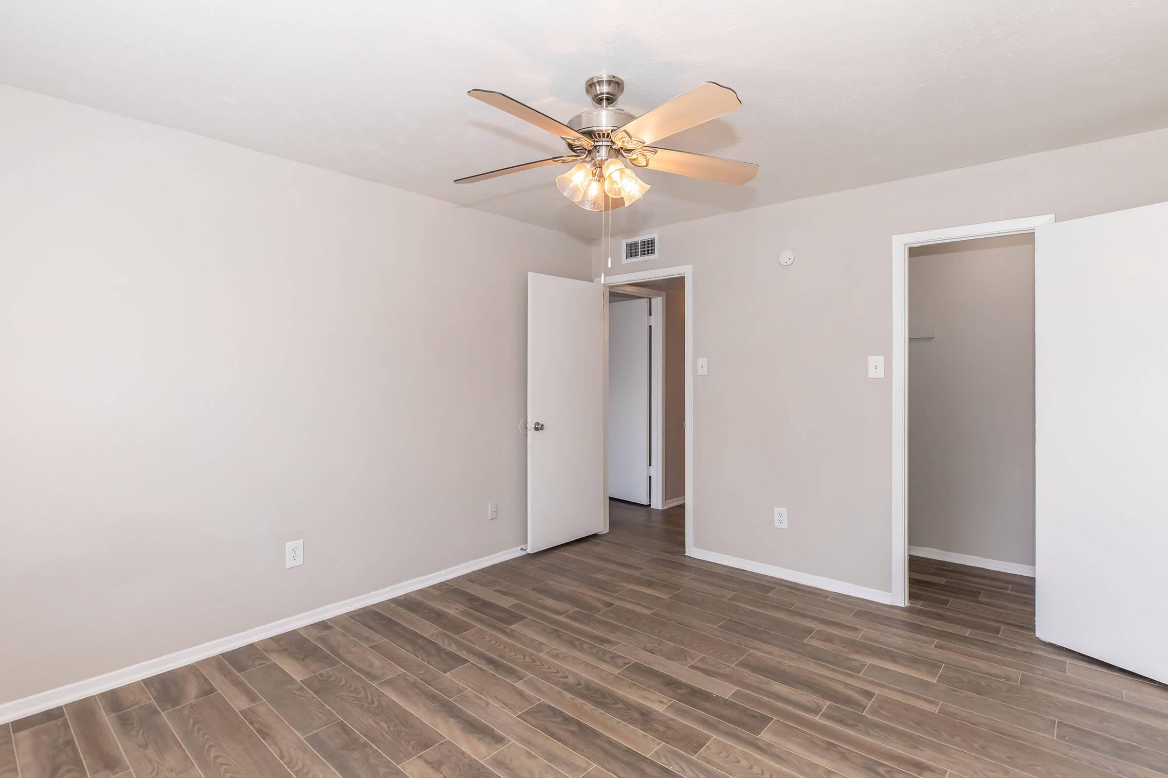 A well-lit, empty room featuring light-colored walls and wooden flooring. It includes a ceiling fan with multiple blades, and two open doors leading to adjacent spaces. The room conveys a clean and spacious atmosphere, suitable for various arrangements.