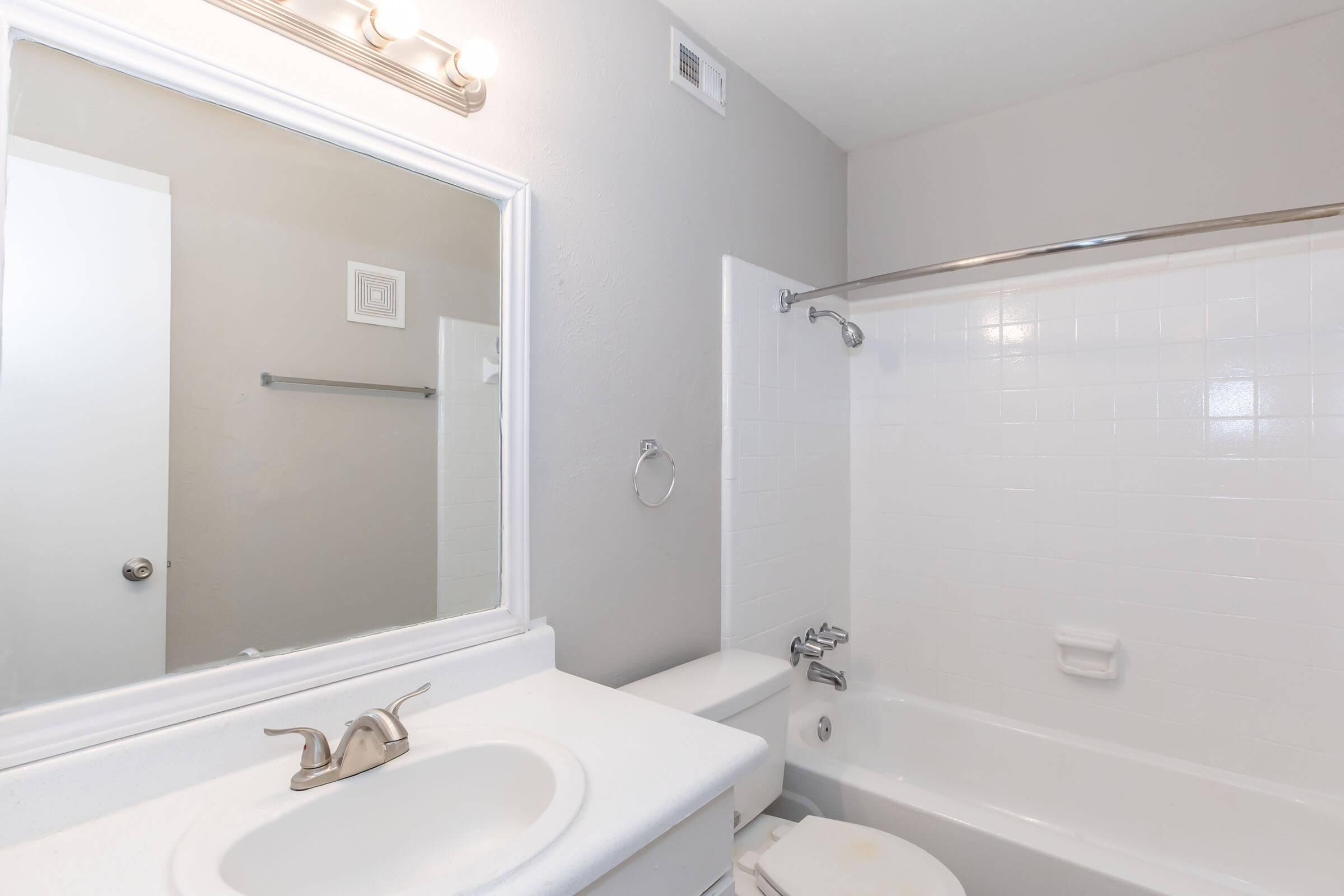 A bathroom featuring a bathtub with a shower, a white sink with a silver faucet, and a large mirror above the sink. The walls are painted light gray, and there is a towel bar and a toilet visible in the space. The overall design is clean and modern.