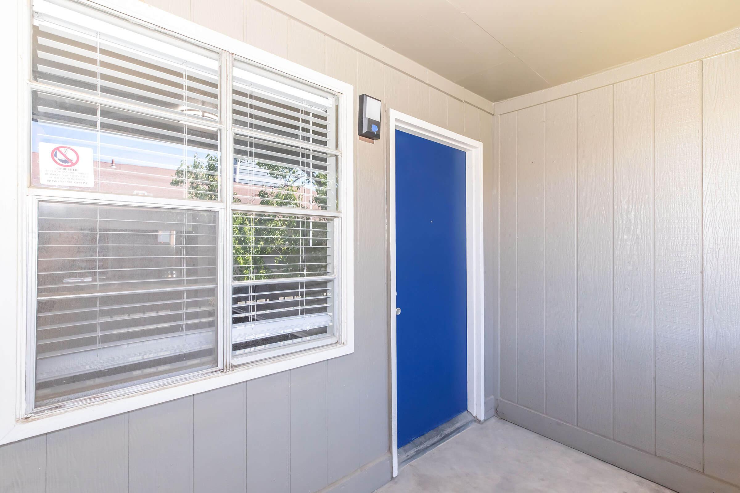 A well-lit entryway featuring a blue door next to a window with blinds. The wall is gray, and there is a small light fixture above the door. A no-smoking sign is visible on the window. The area has a clean, modern appearance with a concrete floor.