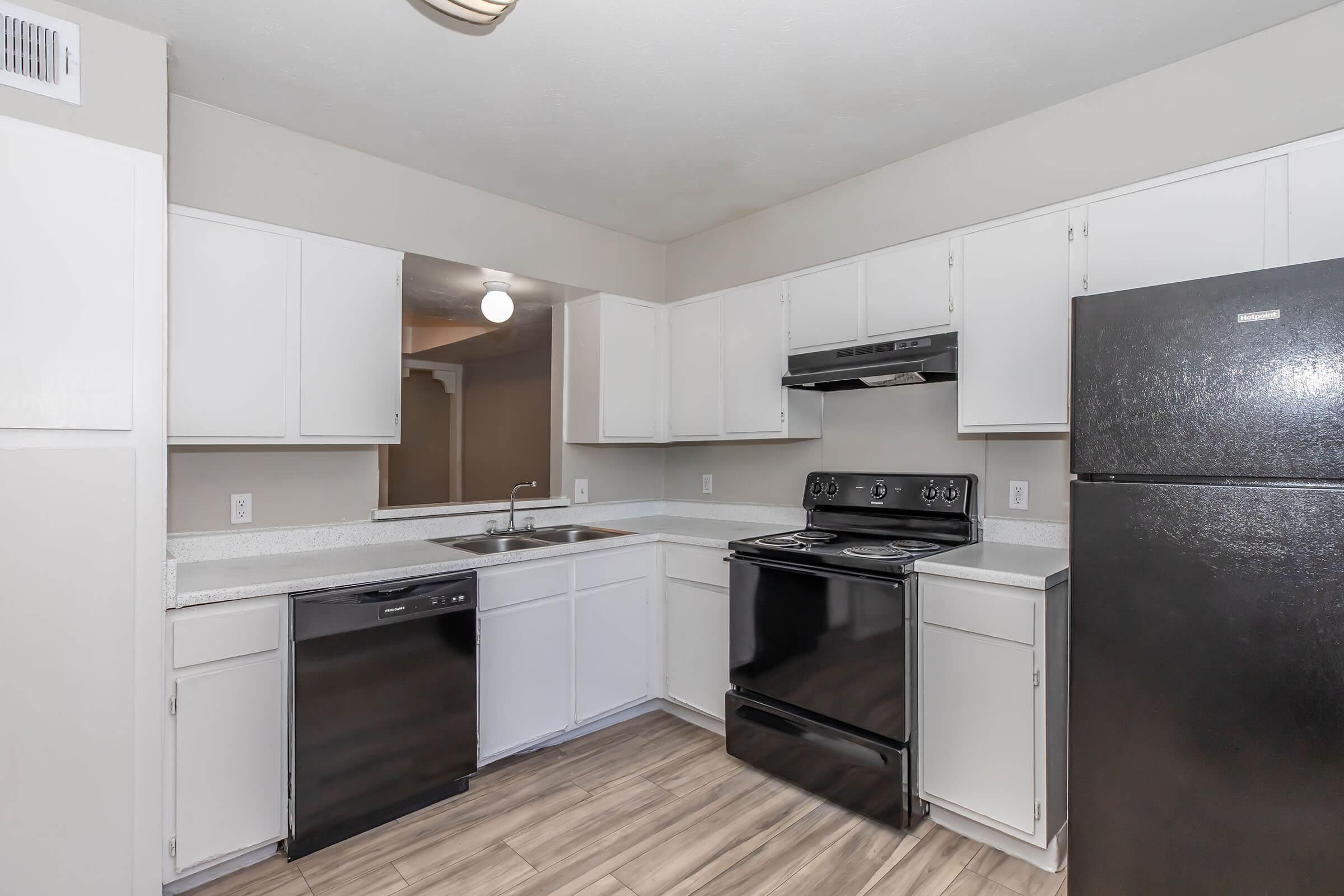 A modern kitchen featuring white cabinetry, a black refrigerator, a black stove, and a dishwasher. The countertop is light-colored, and there is a sink with a faucet. The flooring is a wood-like design. The room is well-lit with a ceiling light fixture.
