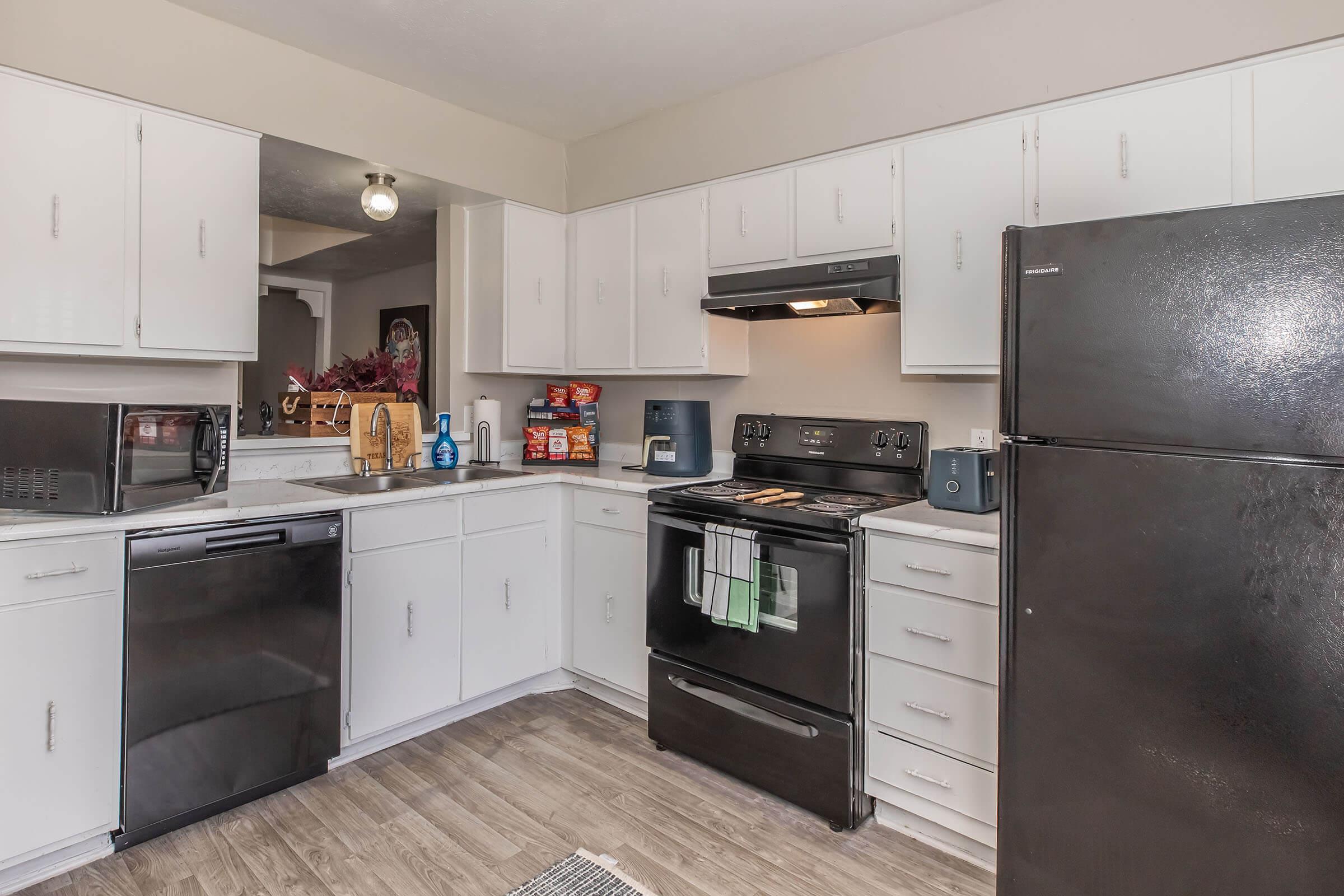 A modern kitchen featuring white cabinets and countertops, a black refrigerator, a black oven, and a black dishwasher. There is a microwave and various kitchen items on the counters, along with a small rug on the floor. The space is bright with overhead lighting, creating an inviting atmosphere.