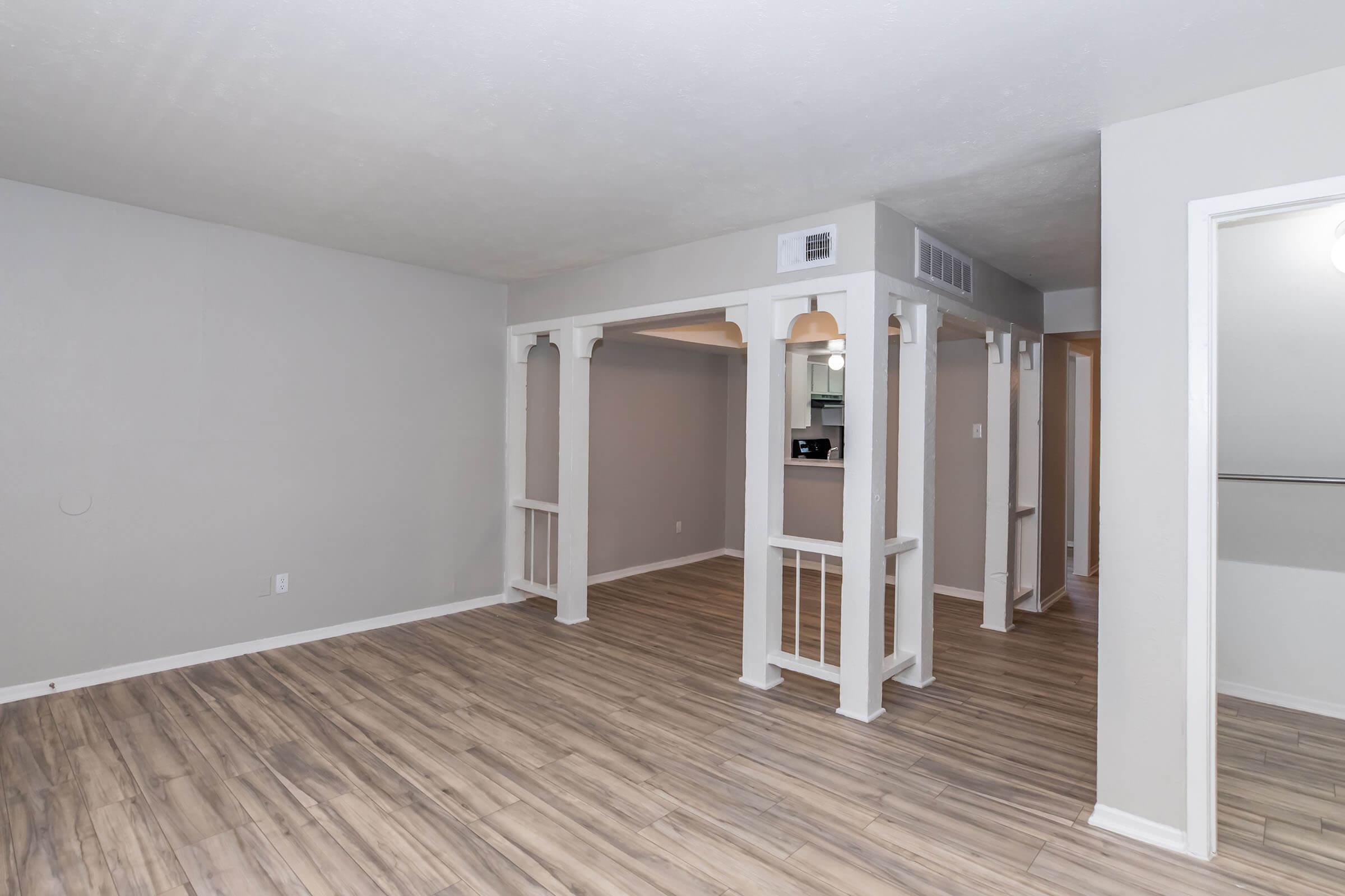 A spacious, unfurnished room with light gray walls and vinyl flooring. It features a distinct architectural structure with white columns and an open layout connecting to adjacent areas. Soft lighting fixtures are visible in the background, creating a welcoming atmosphere.