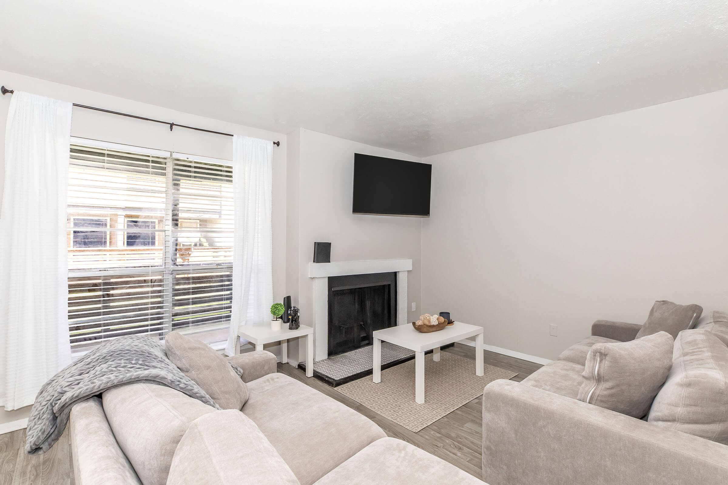 A cozy living room featuring two light gray sofas, a simple white coffee table, and a decorative centerpiece. A window with white curtains lets in natural light, and a flat-screen TV is mounted on the wall above a modern fireplace. The room has neutral-colored walls and a rug on the floor.