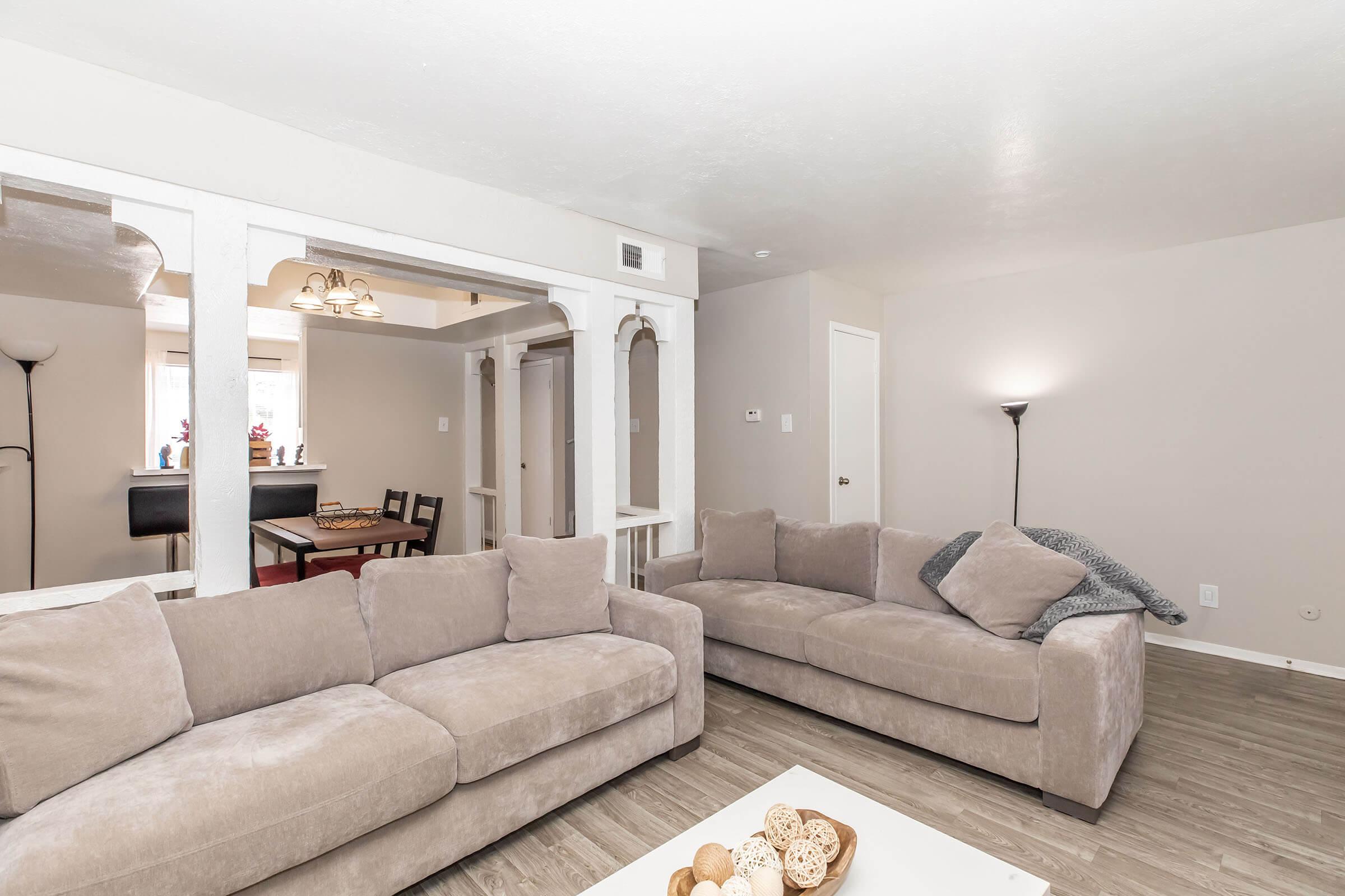 A cozy living room featuring two light grey sofas, a wooden coffee table with decorative items, and an open layout connecting to a dining area. The walls are painted in a soft neutral color, and there is a standing lamp for additional lighting in the space.