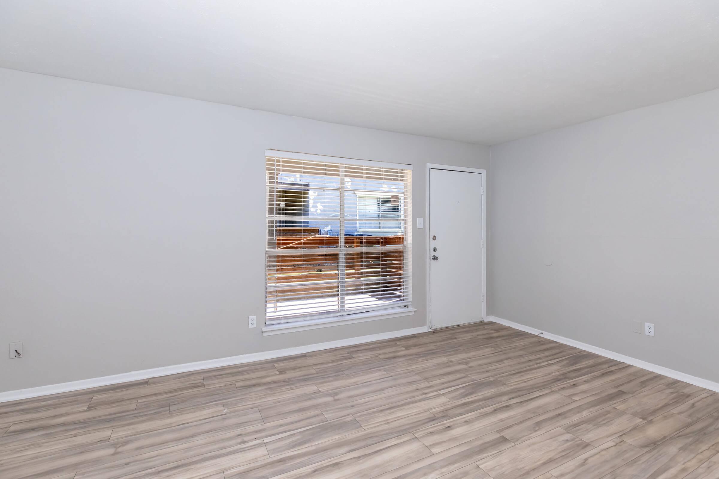 Empty room with light gray walls and laminate flooring. A large window with white blinds allows natural light, and there is a door on the right leading outside. The space appears clean and modern, ideal for furnishing.