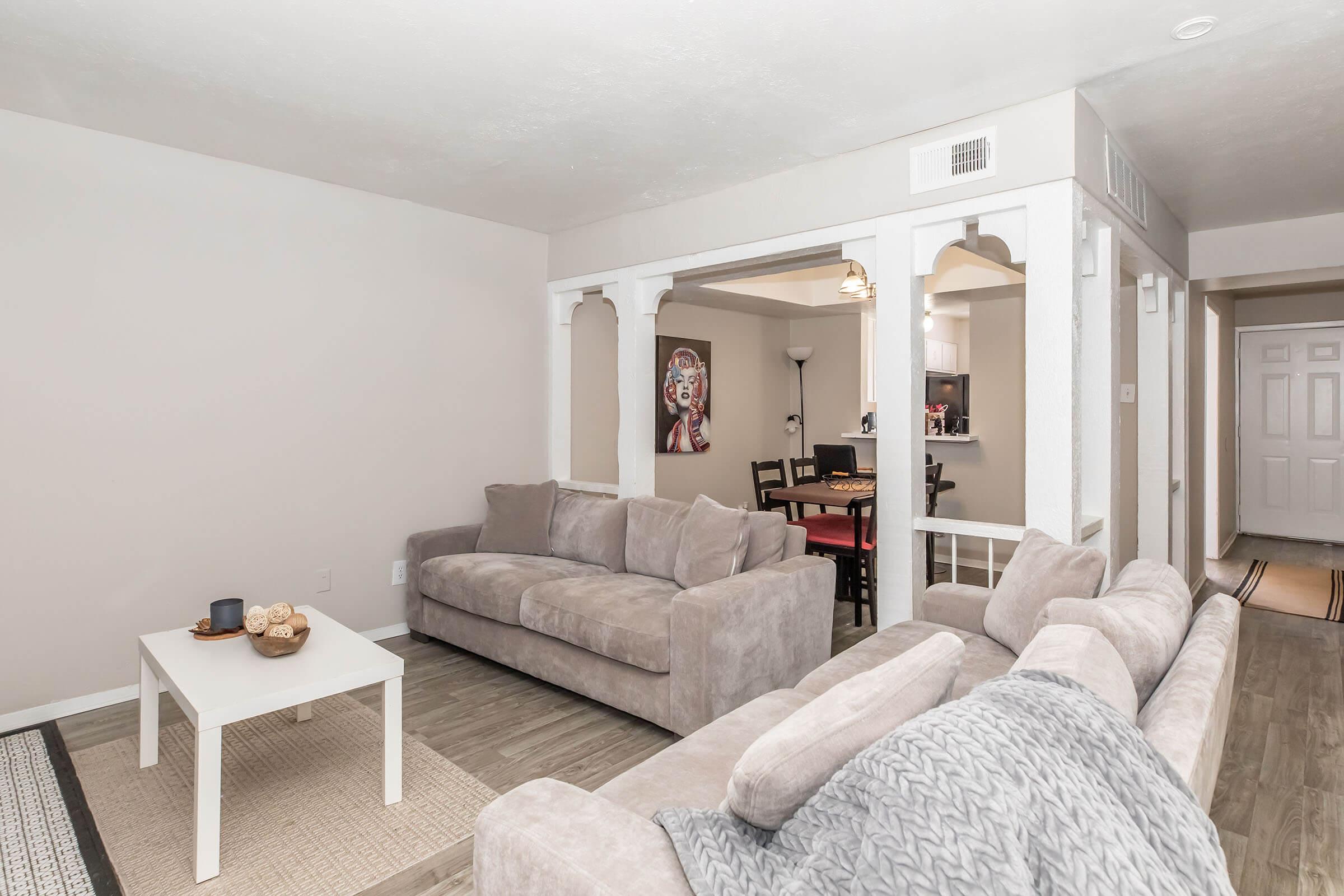 A cozy living room featuring a light gray sectional sofa and a small white coffee table. The walls are painted a soft neutral color, and there's a textured area rug on the floor. In the background, a dining area with a black table and chairs is visible, along with a glimpse of a kitchen.