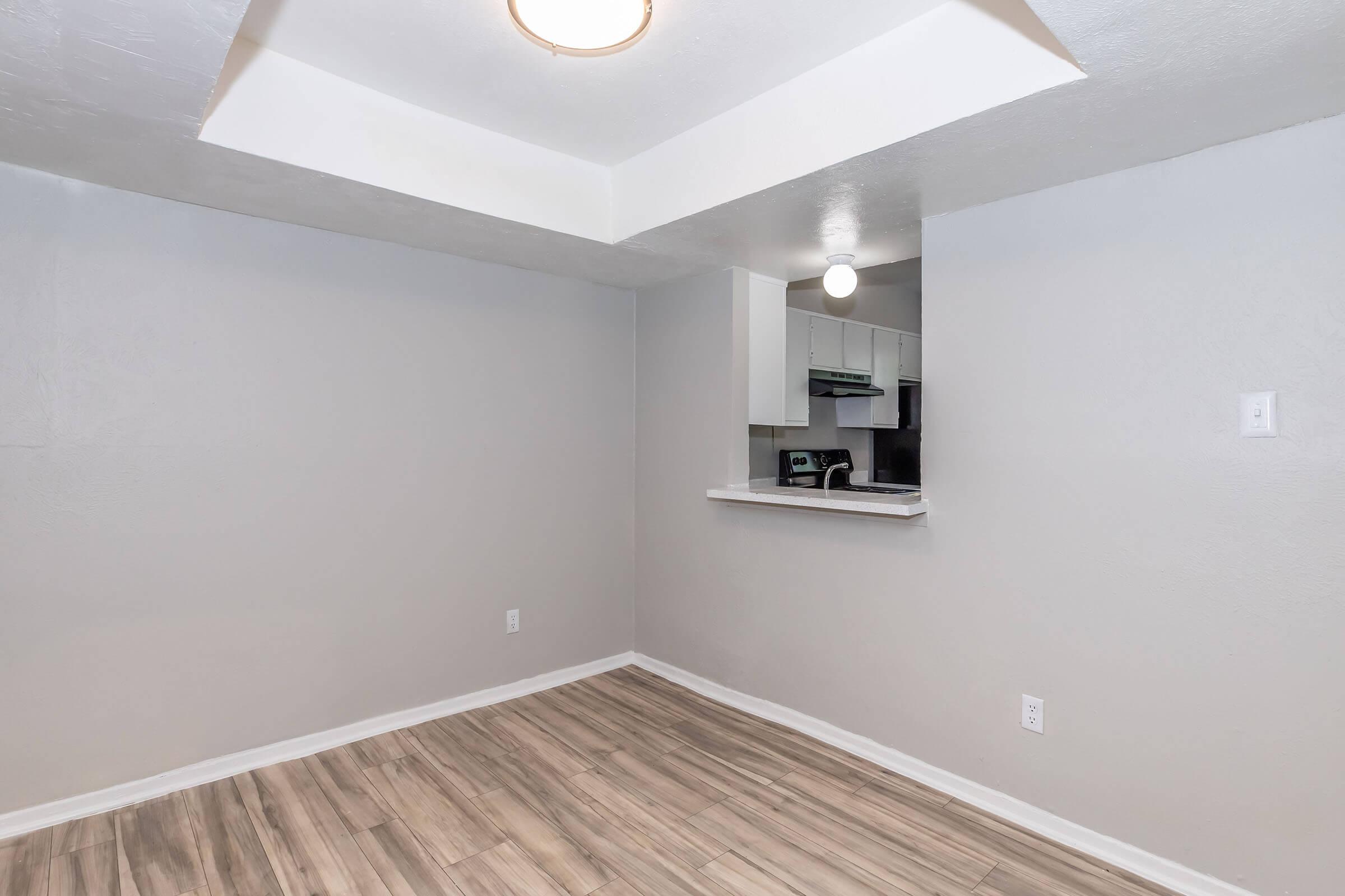 A spacious, empty room with light gray walls and a wood-like floor. A small kitchen area is visible in the background, featuring a counter and cabinets. The ceiling has a modern light fixture, and the overall atmosphere is bright and inviting.