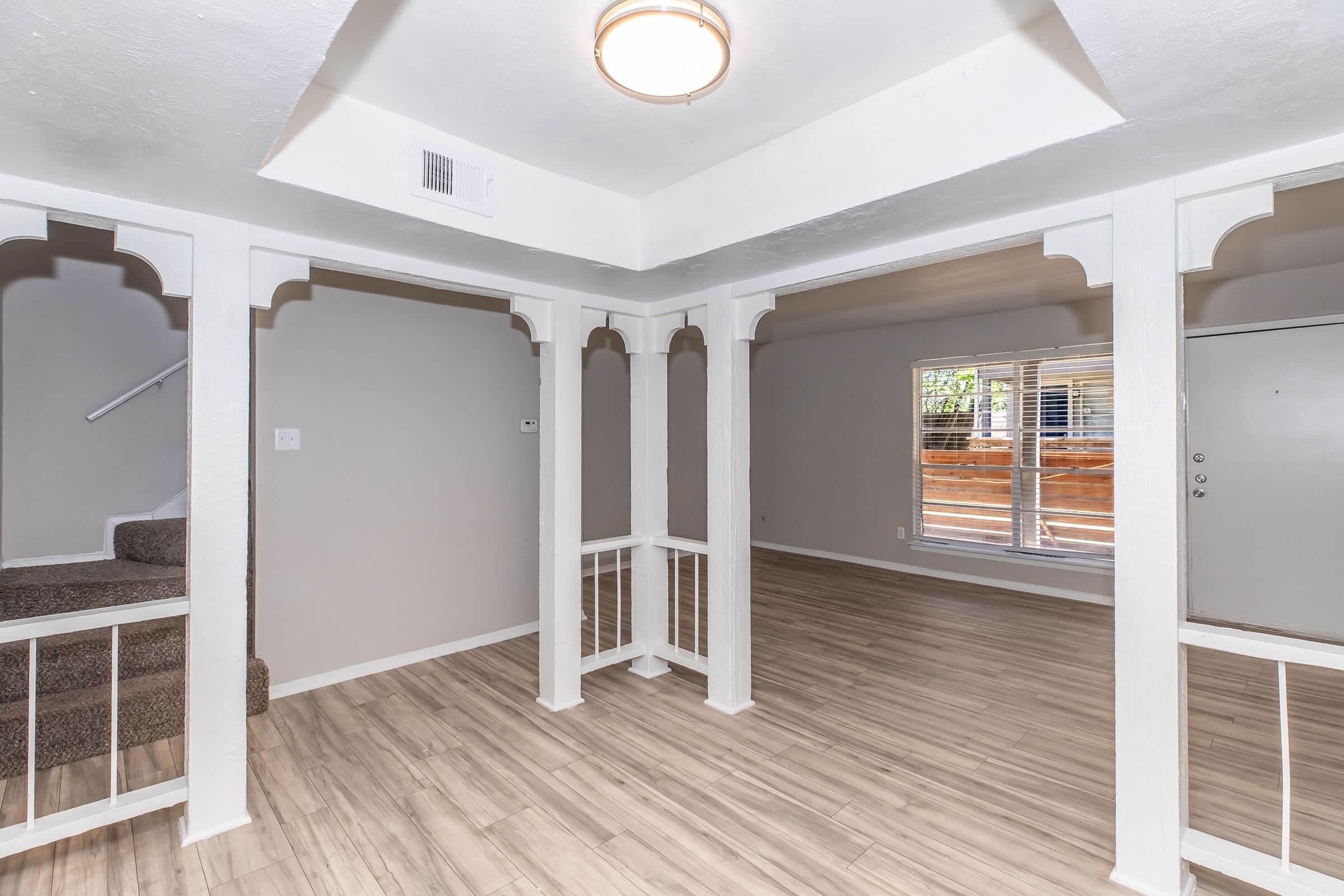 Interior view of a spacious living area featuring light-colored walls, a wood-like floor, decorative archways, and large windows providing natural light. A staircase is seen in the background, and the entrance is visible on the right side. The space is uncluttered, creating an open and inviting atmosphere.